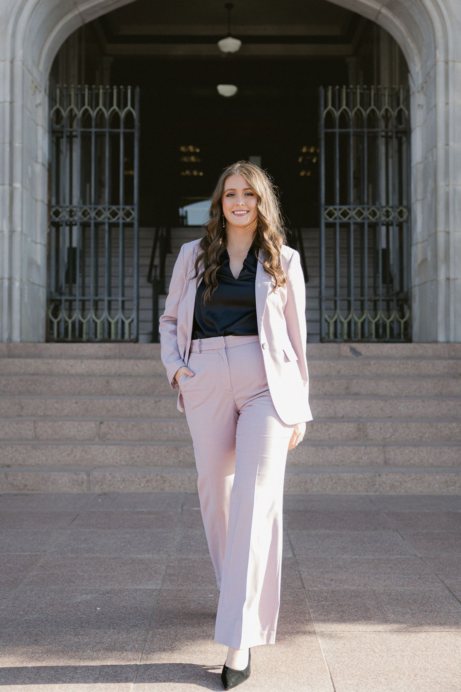 Professional woman in light pink pantsuit and white blazer poses near ornate building entrance in five different poses.