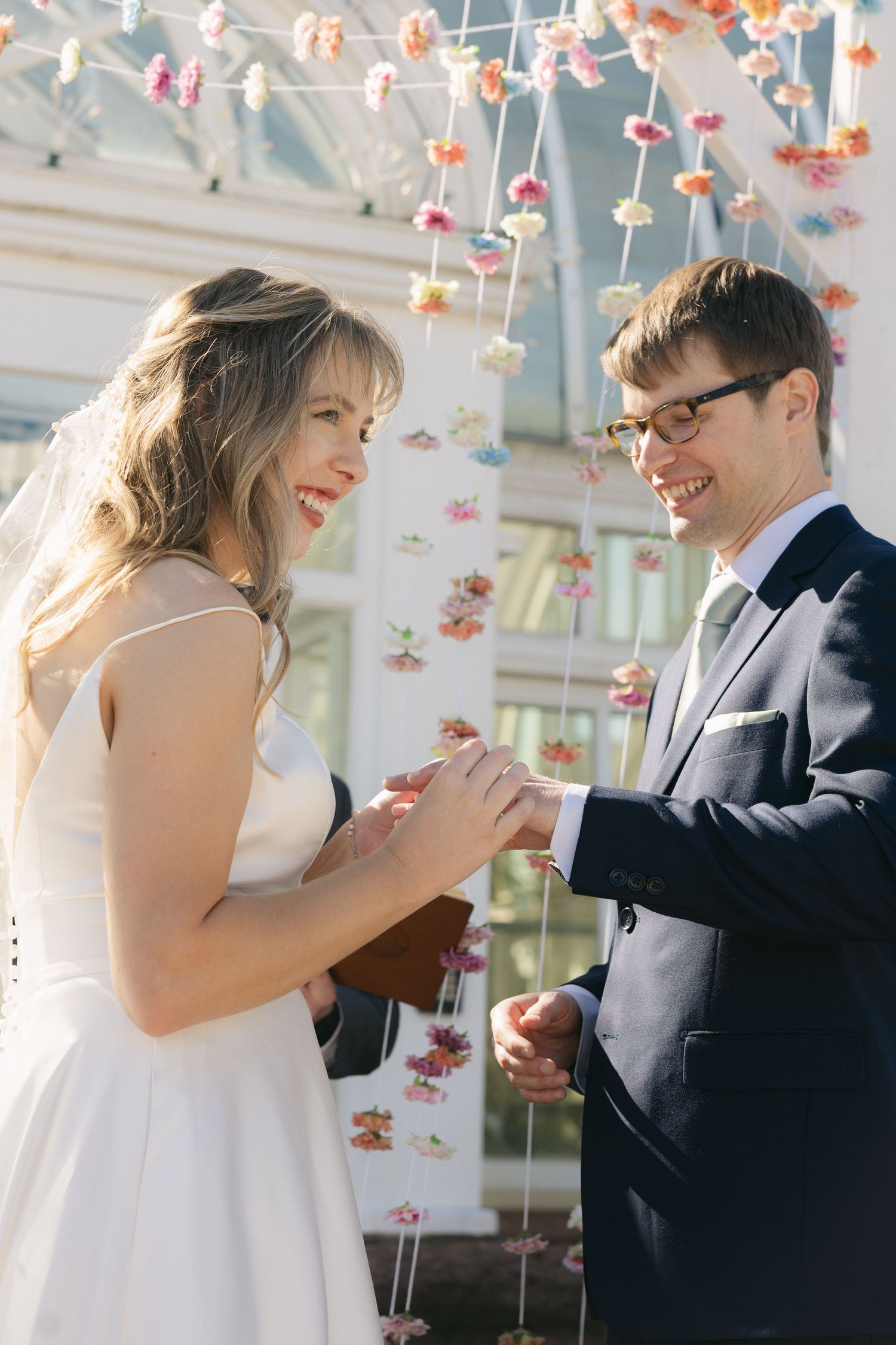 Bride and groom exchanging rings at outdoor wedding ceremony with colorful hanging floral decorations.