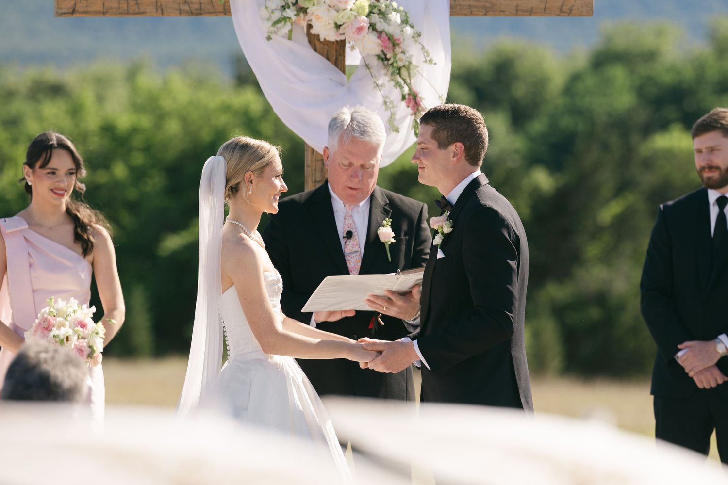 Bride and groom exchange vows outdoors at a romantic wedding ceremony with floral arch and lush green hills.