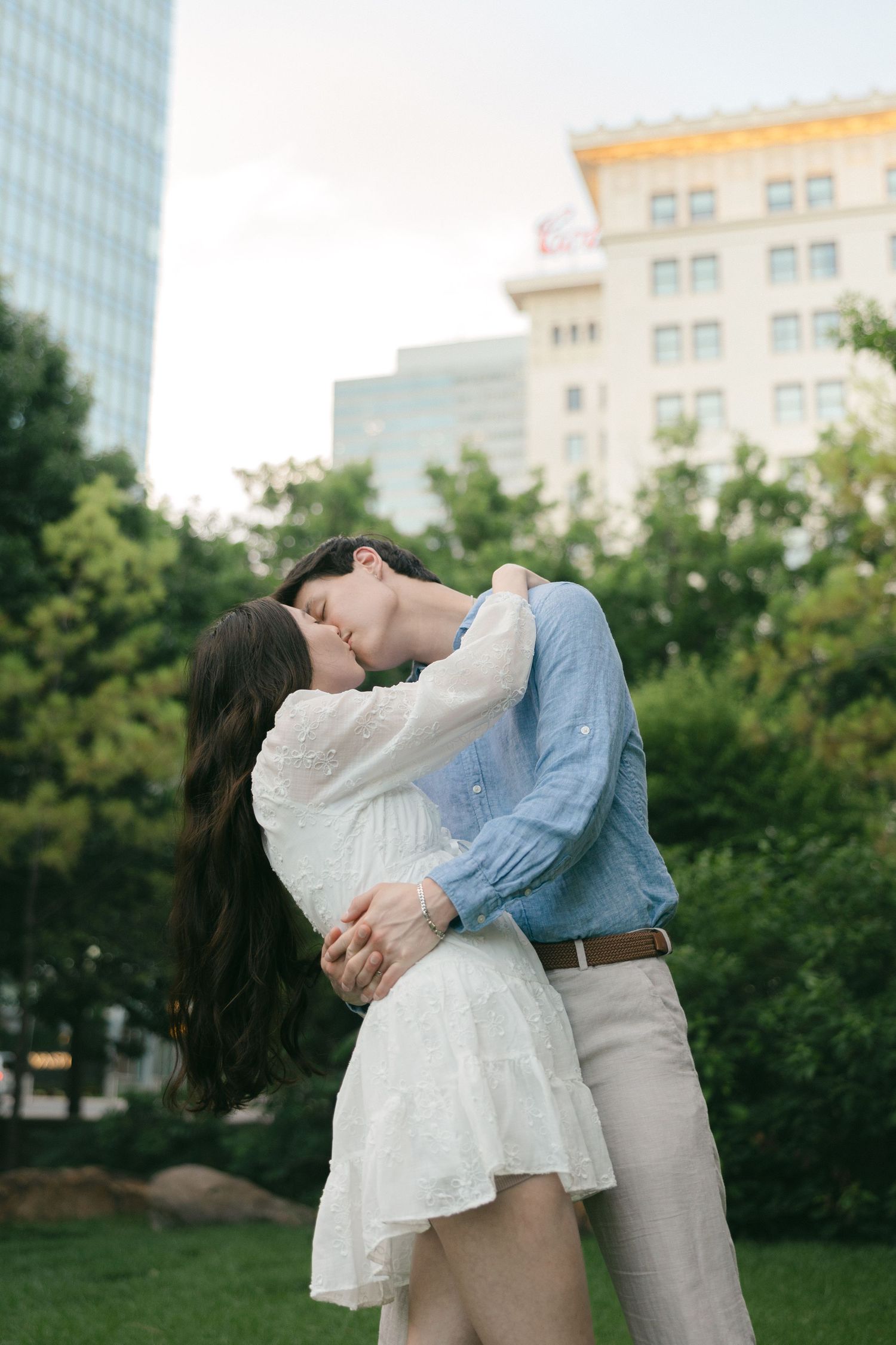 A couple shares a romantic kiss in a lush urban park, woman in white dress embraced by man in blue shirt.