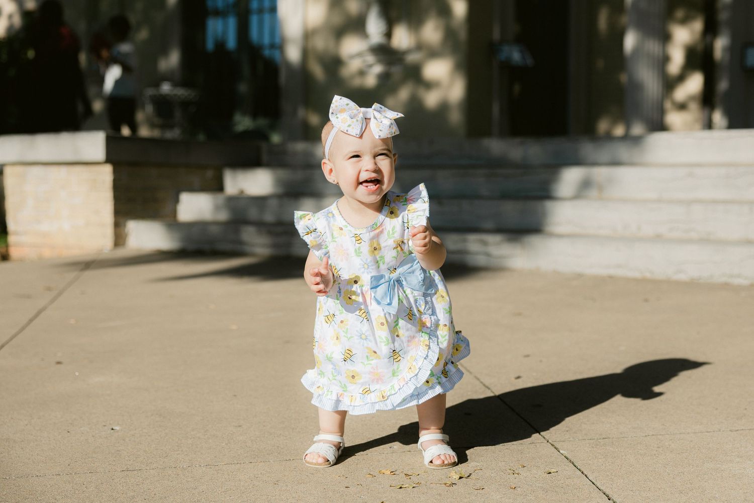 A small child in a floral dress and bow stands on sunny stone steps casting a shadow.