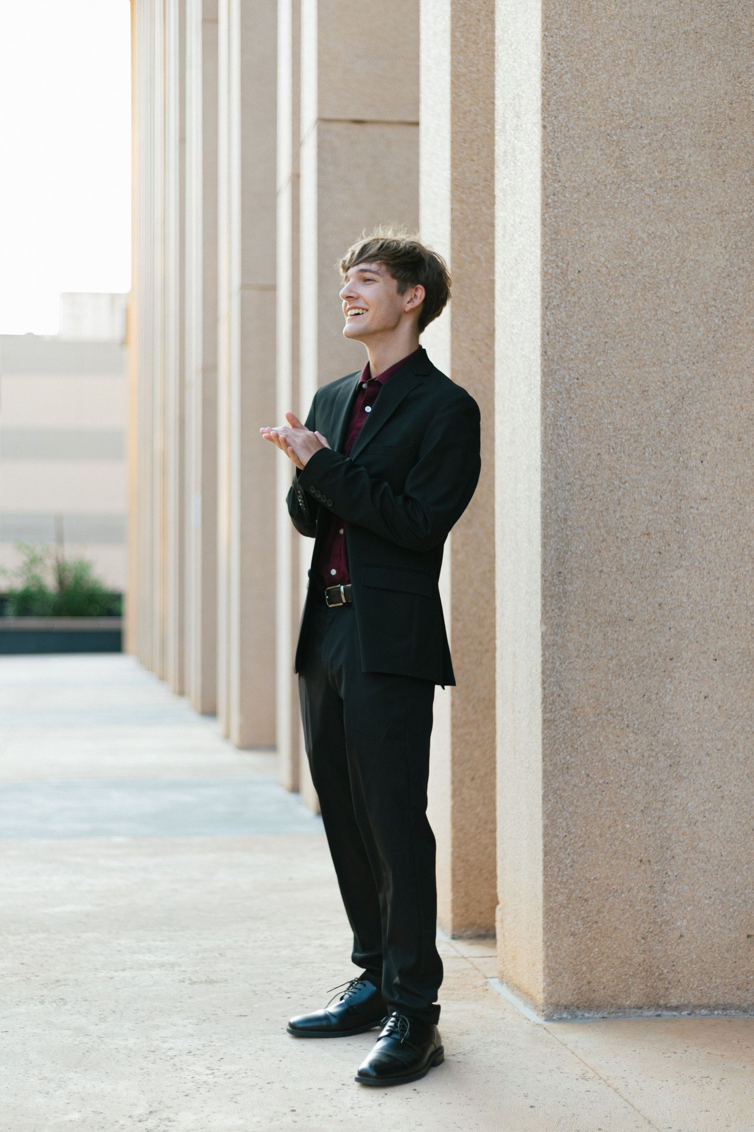 Young person in formal black suit laughing while standing against beige columns.
