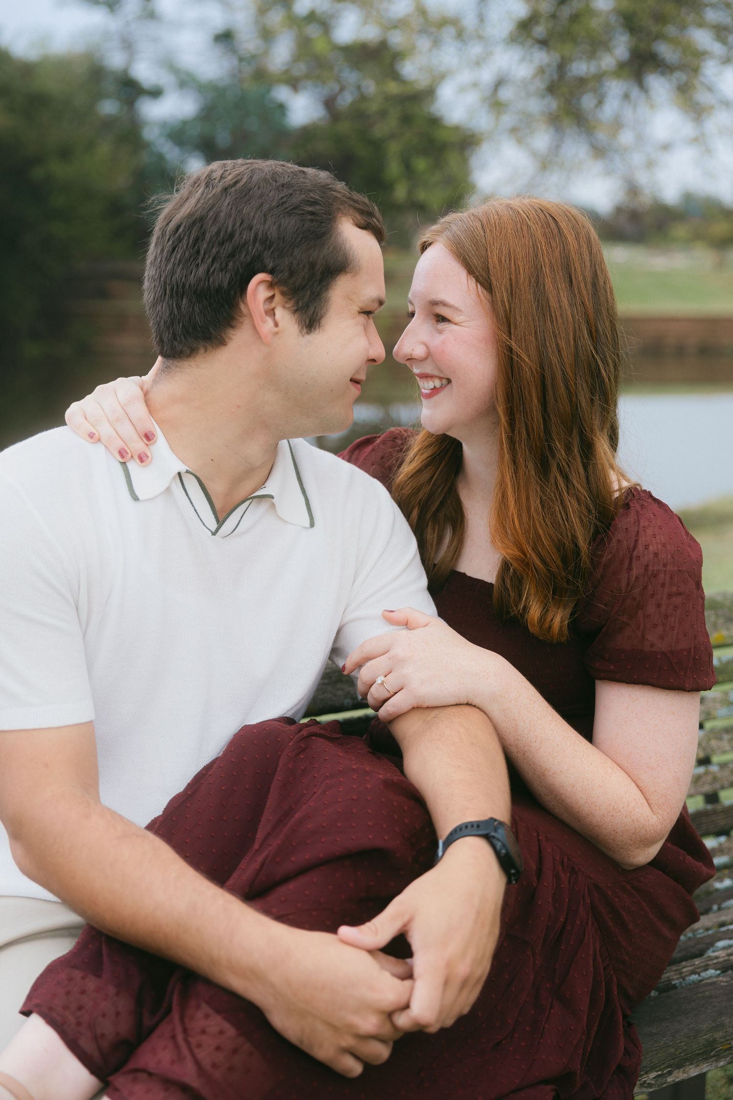 A romantic couple shares an intimate moment by a lake, with her wearing burgundy and him in white.