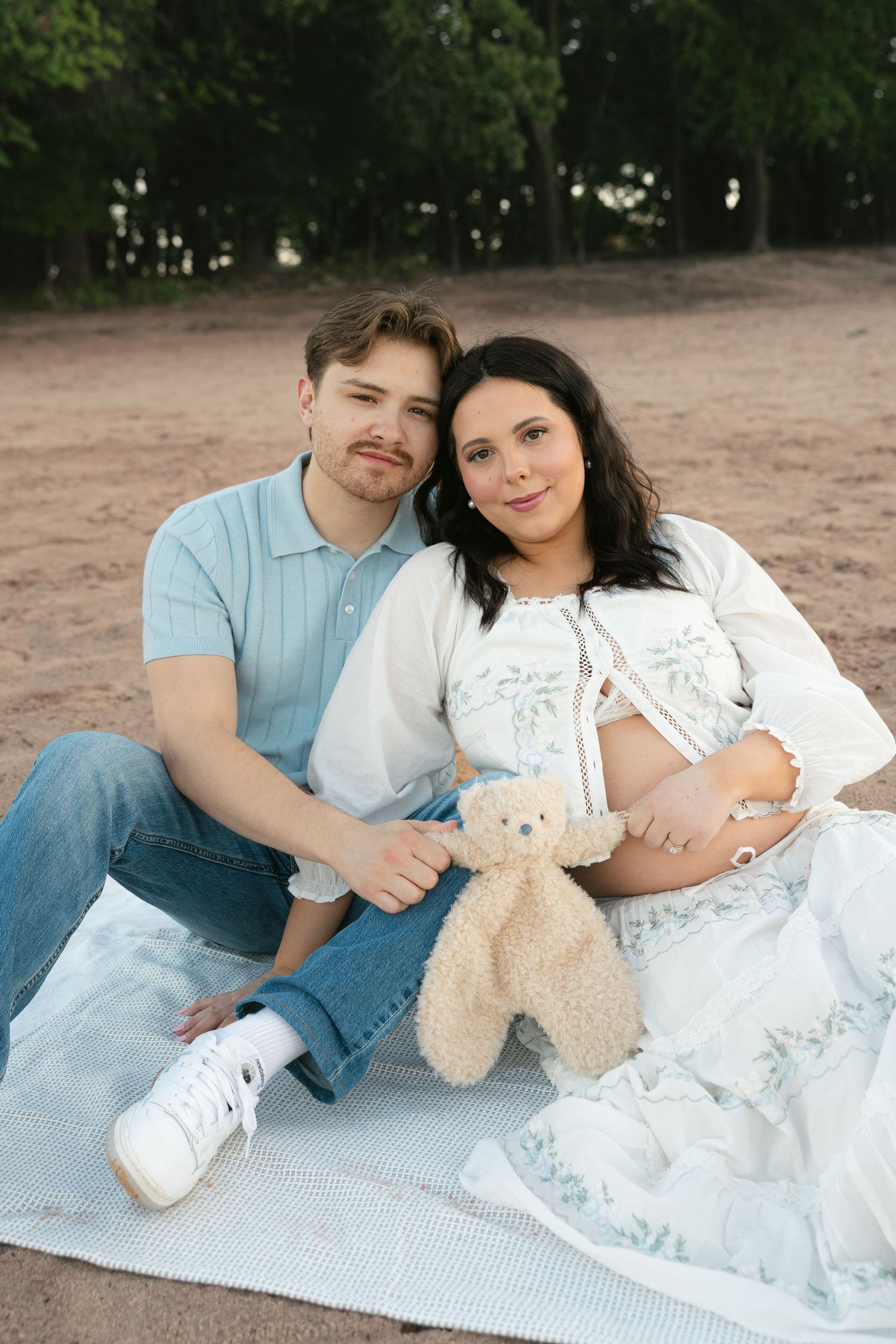 Pregnant couple sitting on blanket at beach, holding teddy bear, smiling at camera, maternity photo shoot outdoors.