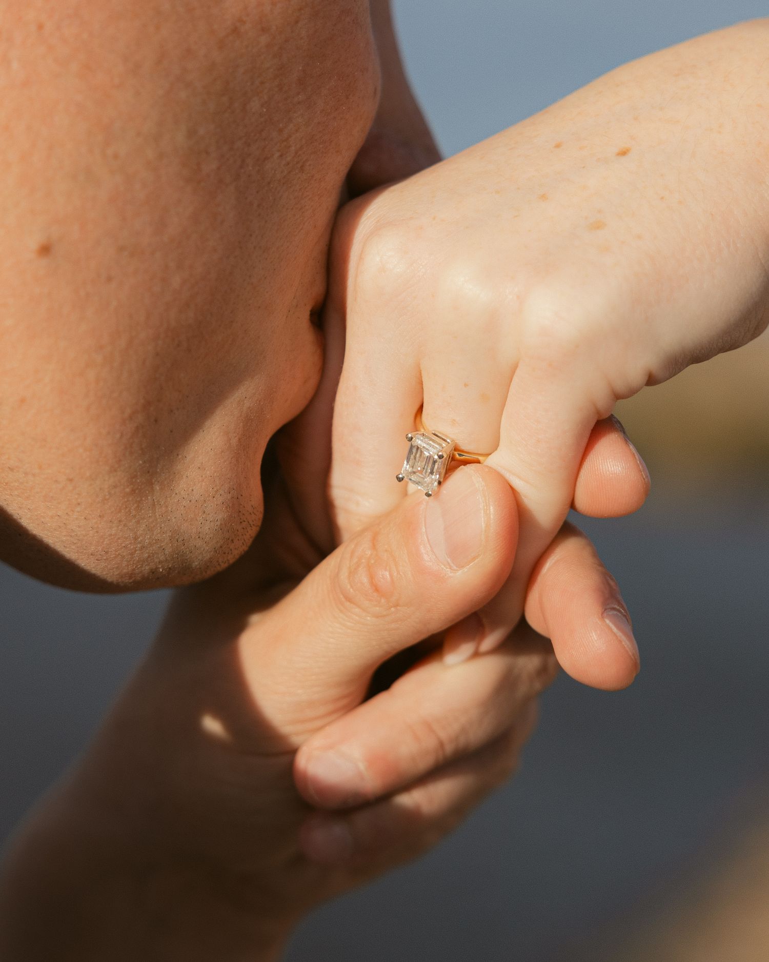 Close-up of someone kissing a hand wearing a diamond engagement ring in soft natural lighting.