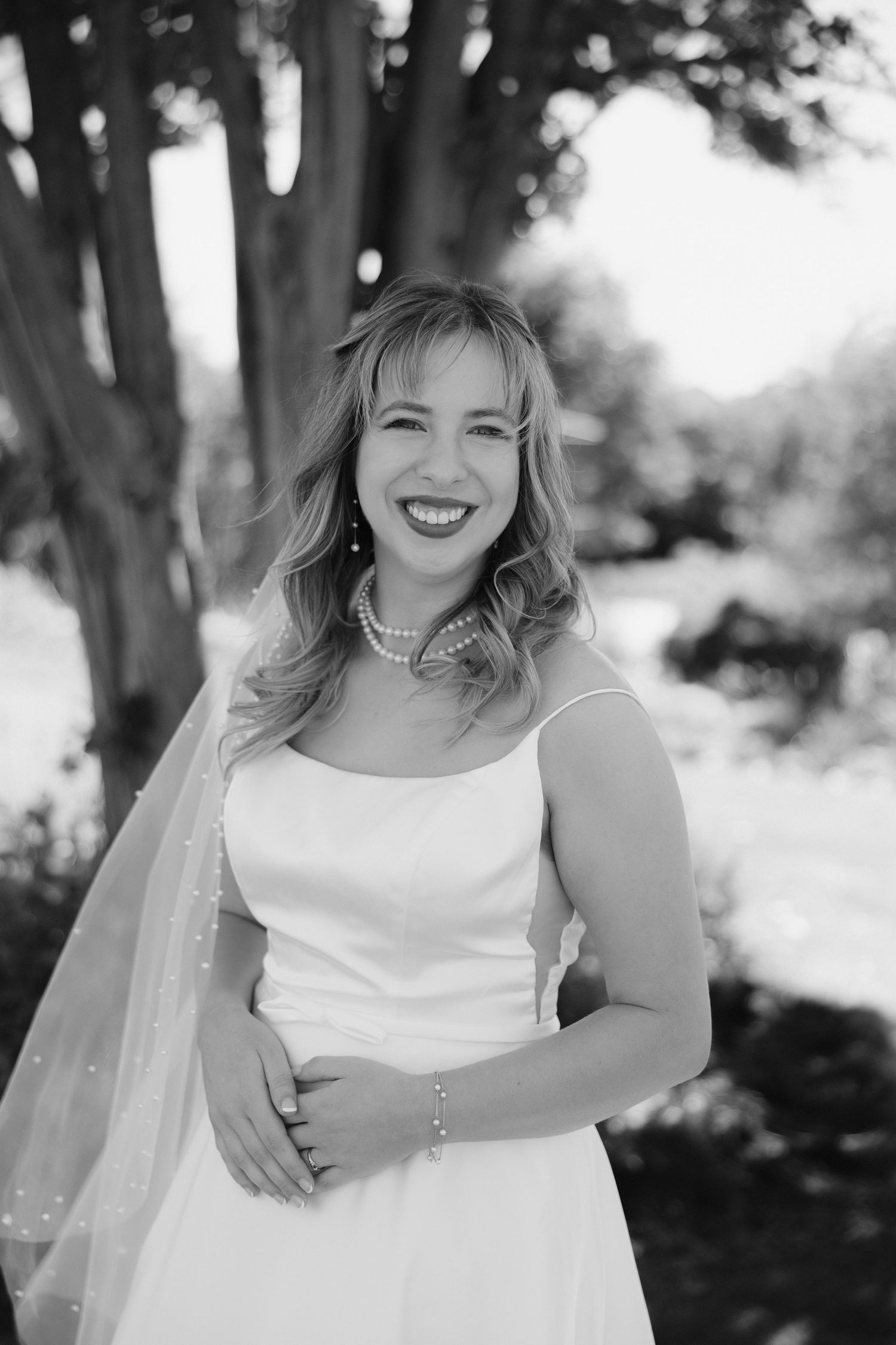 Smiling bride in white gown and veil with pearl jewelry poses outdoors in black and white wedding portrait.