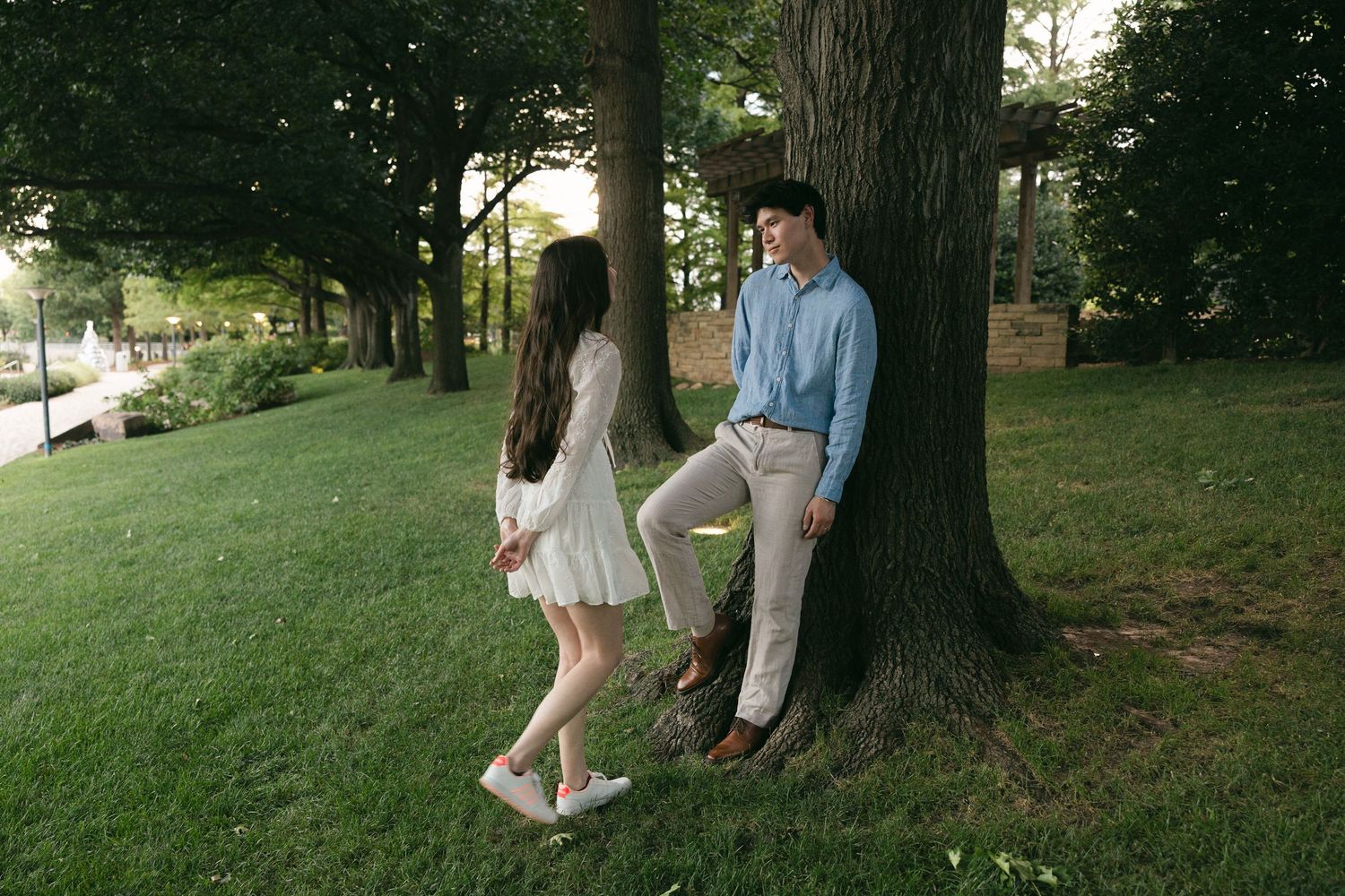 A couple shares a romantic moment in a lush green park, man leaning against a tree in blue shirt and khakis.