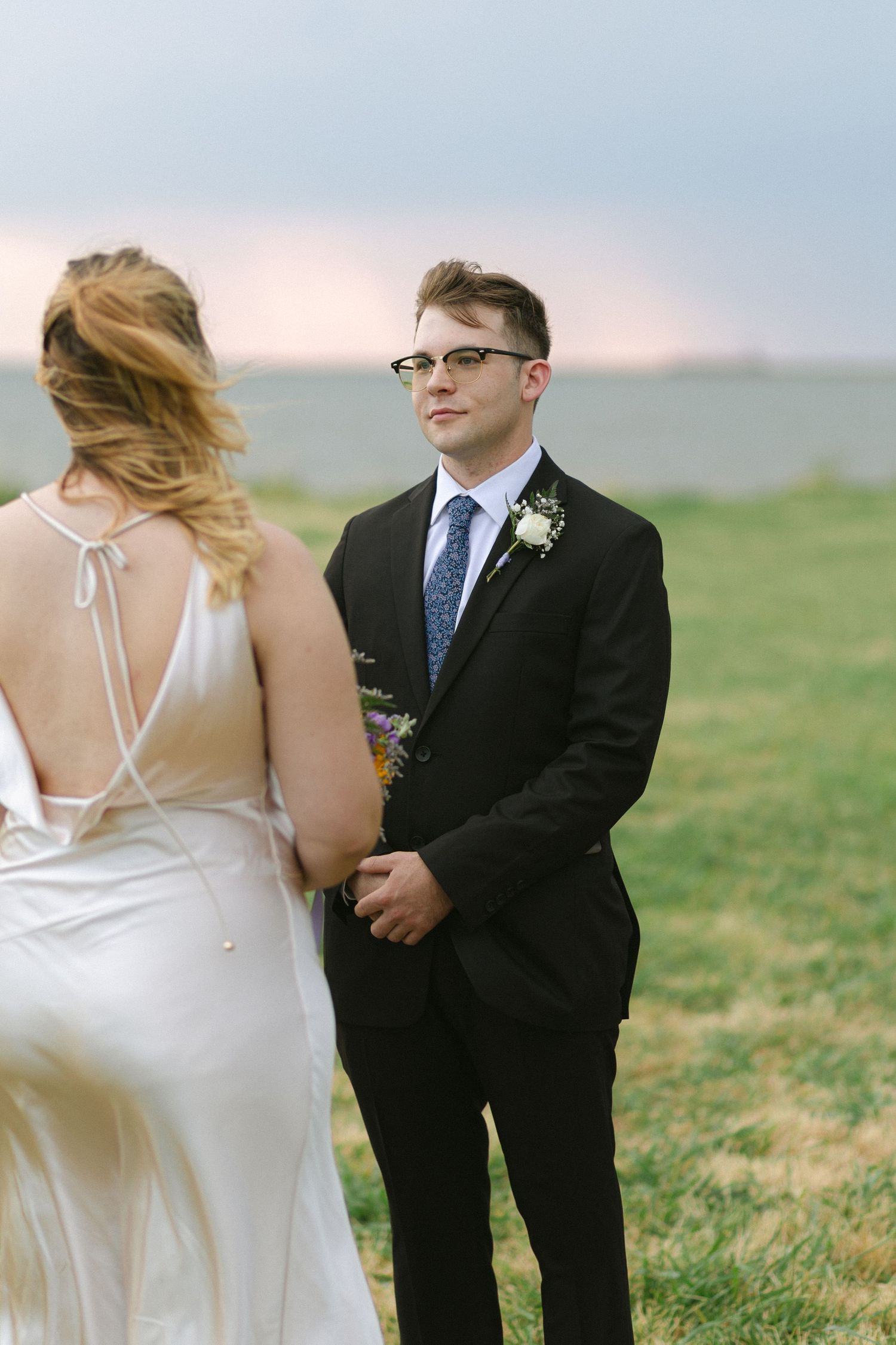 Couple in wedding attire standing together on a grassy hillside at sunset with the bride in white and groom in black suit.