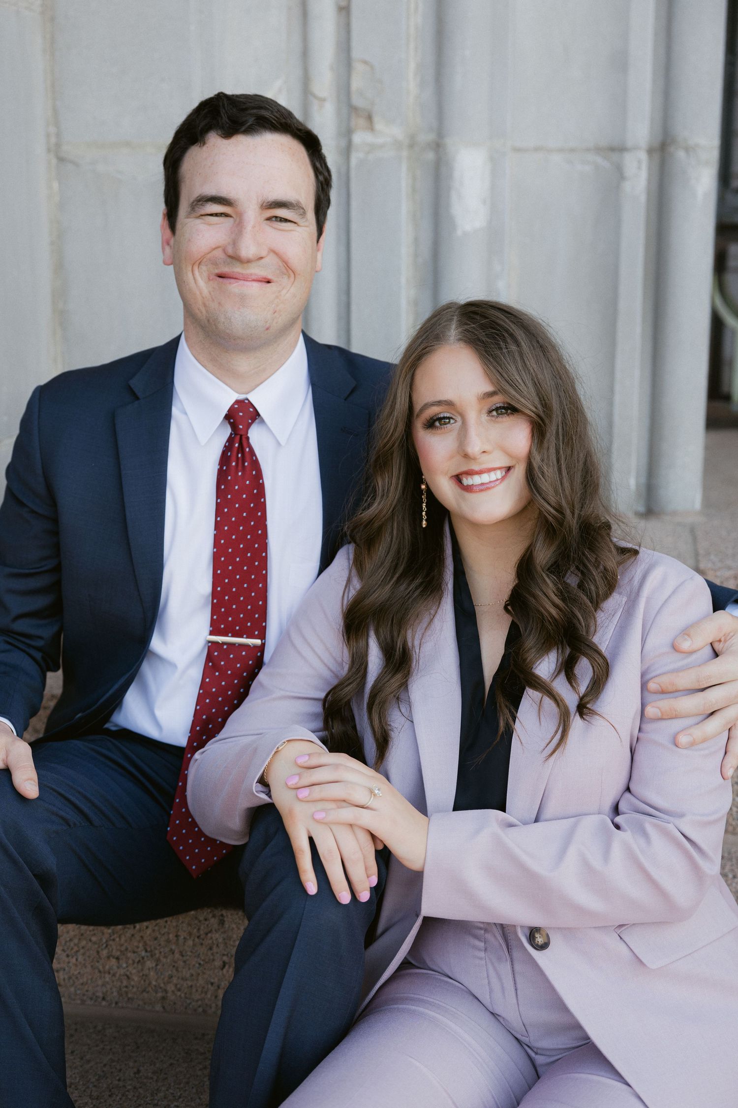A professional couple in business attire sits together and smiles, wearing navy and pink suits with a red polka dot tie.