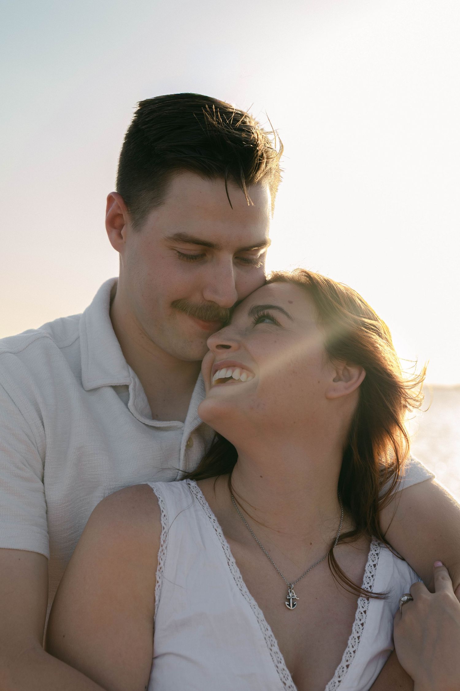A happy couple shares a tender moment outdoors, with the woman smiling up at the man in golden backlit sunlight.