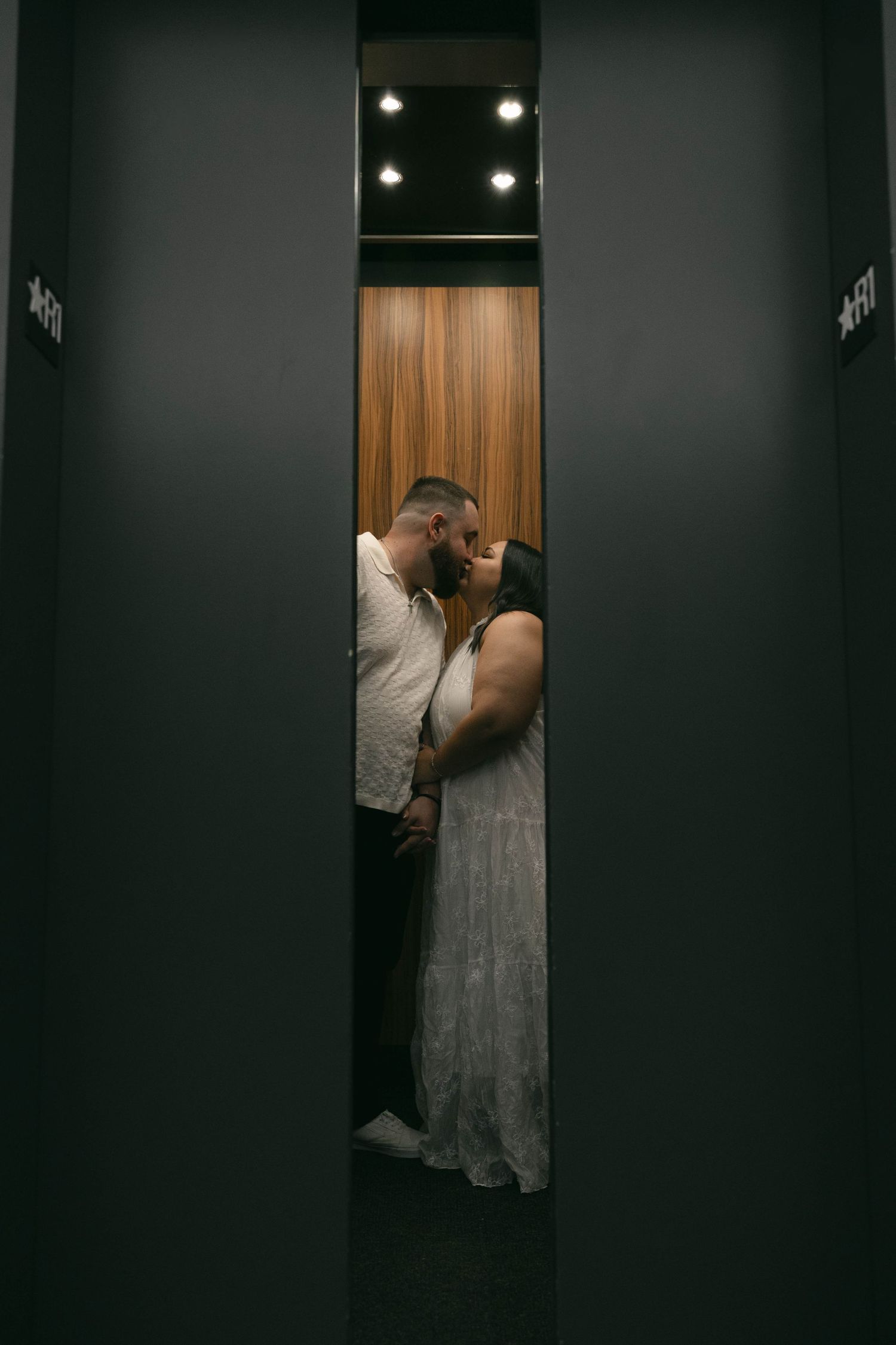 Couple sharing a romantic kiss inside a modern elevator with warm wood paneling and overhead lights.