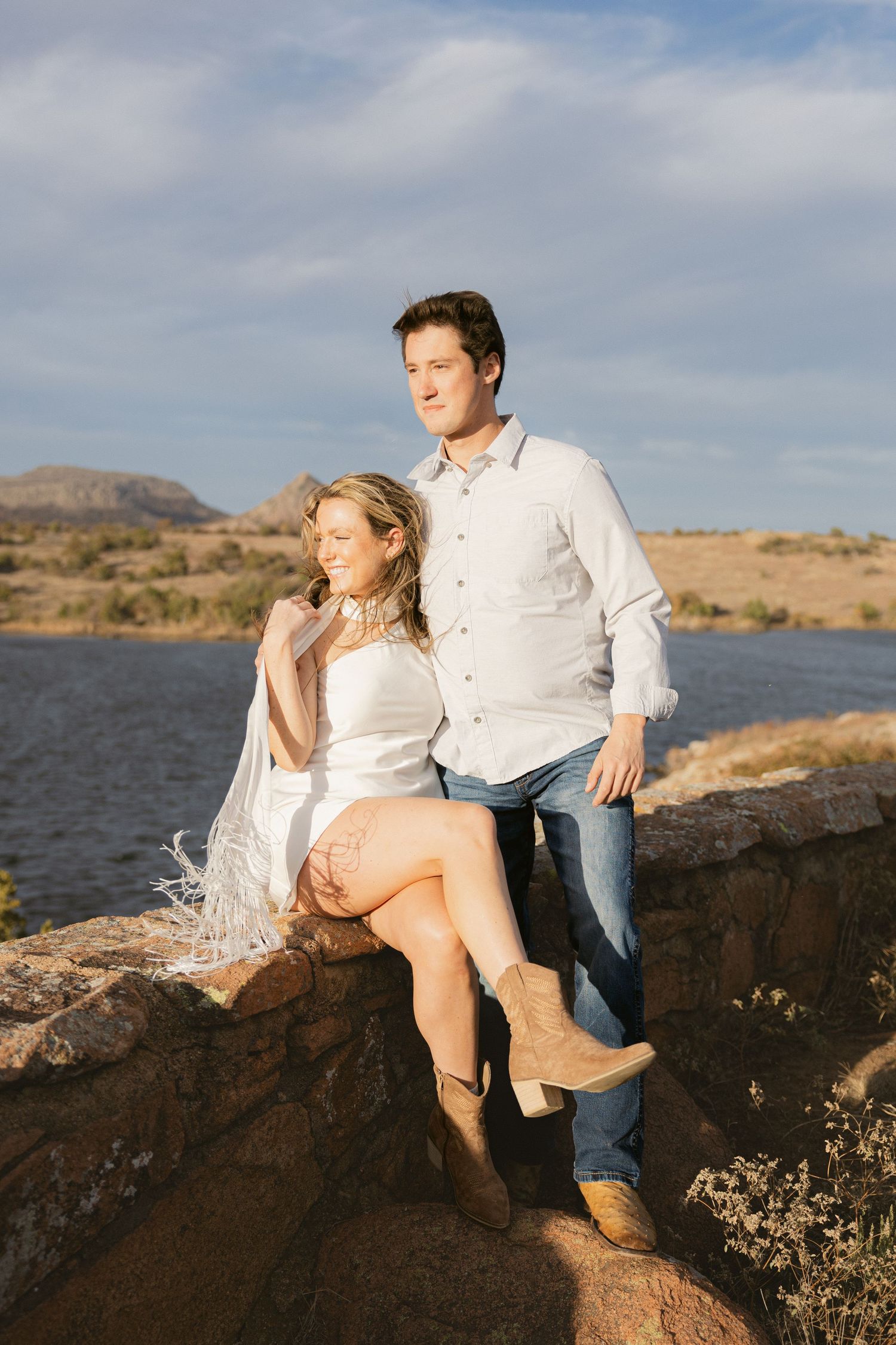 A couple sits together on rocky cliffs overlooking a serene lake during a cloudy afternoon.