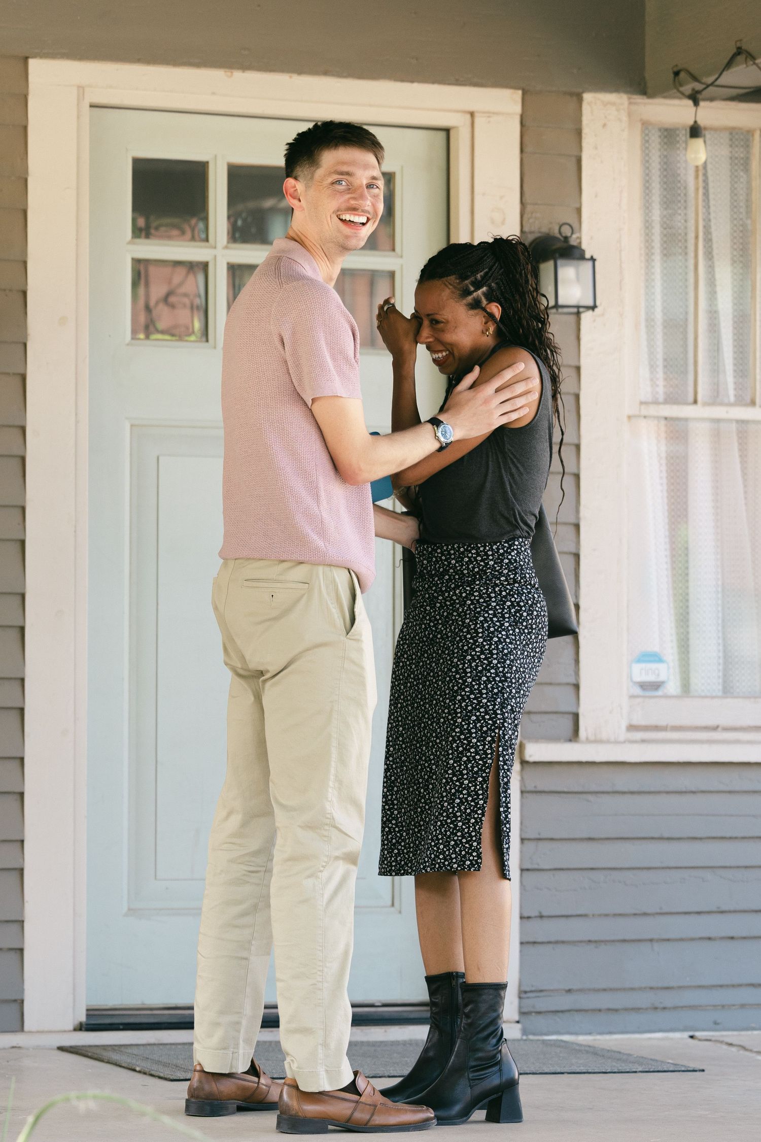 Interracial couple laughing and embracing on a home's front porch, man in pink polo and woman in black dress.