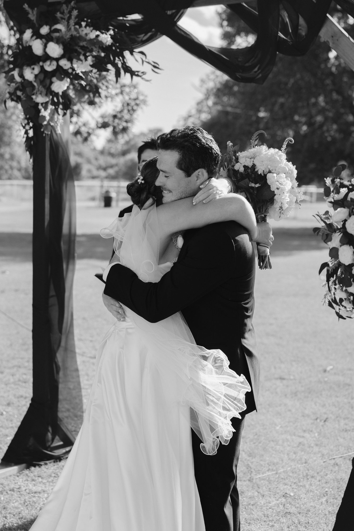 Bride and groom share an emotional embrace under a floral arch during their outdoor wedding ceremony.