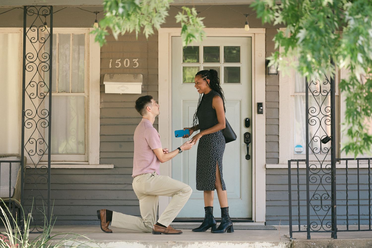 Man kneeling on porch proposing to woman outside house number 1503 on a sunny day.