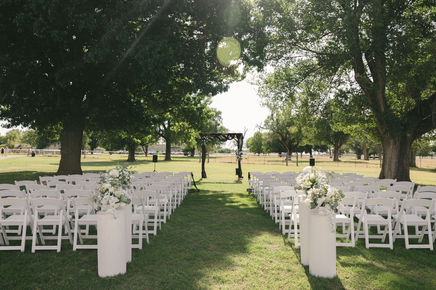 Outdoor wedding ceremony setup with white folding chairs, floral arrangements, and a black arch under large shade trees on green grass.