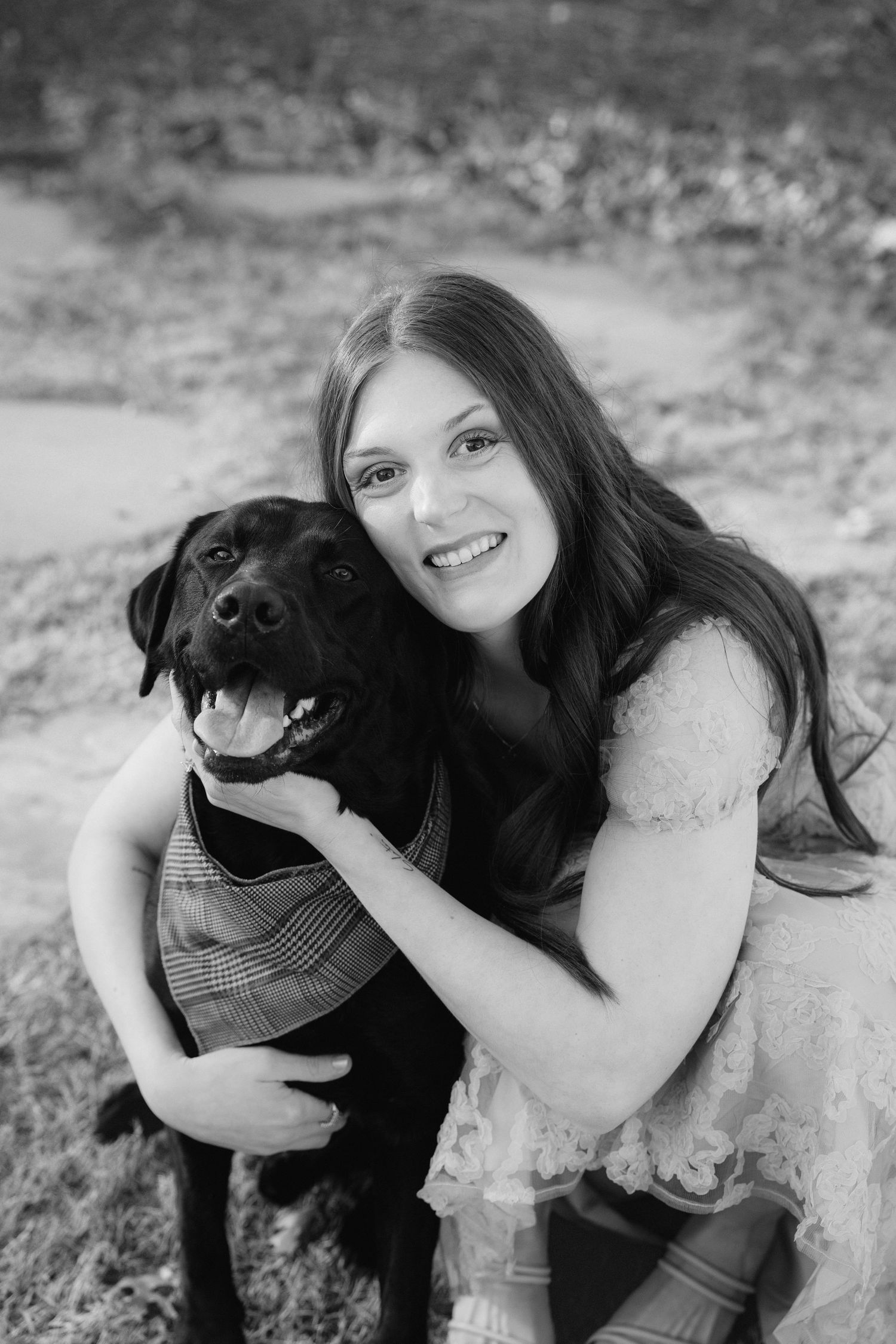 A happy black labrador dog wearing a plaid vest poses with their companion in a black and white outdoor portrait.