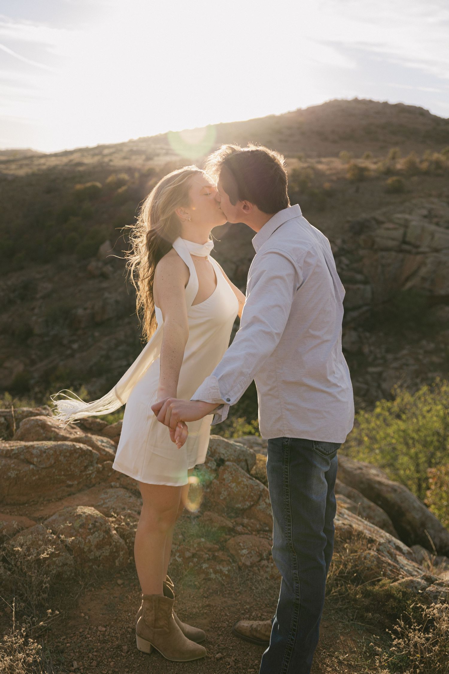 A romantic couple sharing a kiss at sunset on a rocky hillside with mountains in the background.