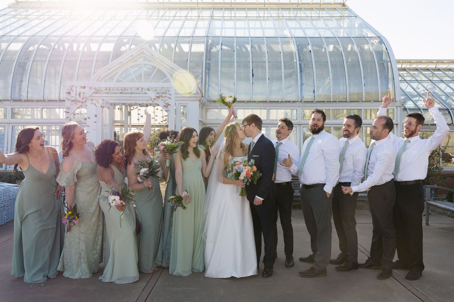 Wedding party poses outside a glass greenhouse, bridesmaids in sage green gowns, groomsmen in gray suits, couple kissing center.