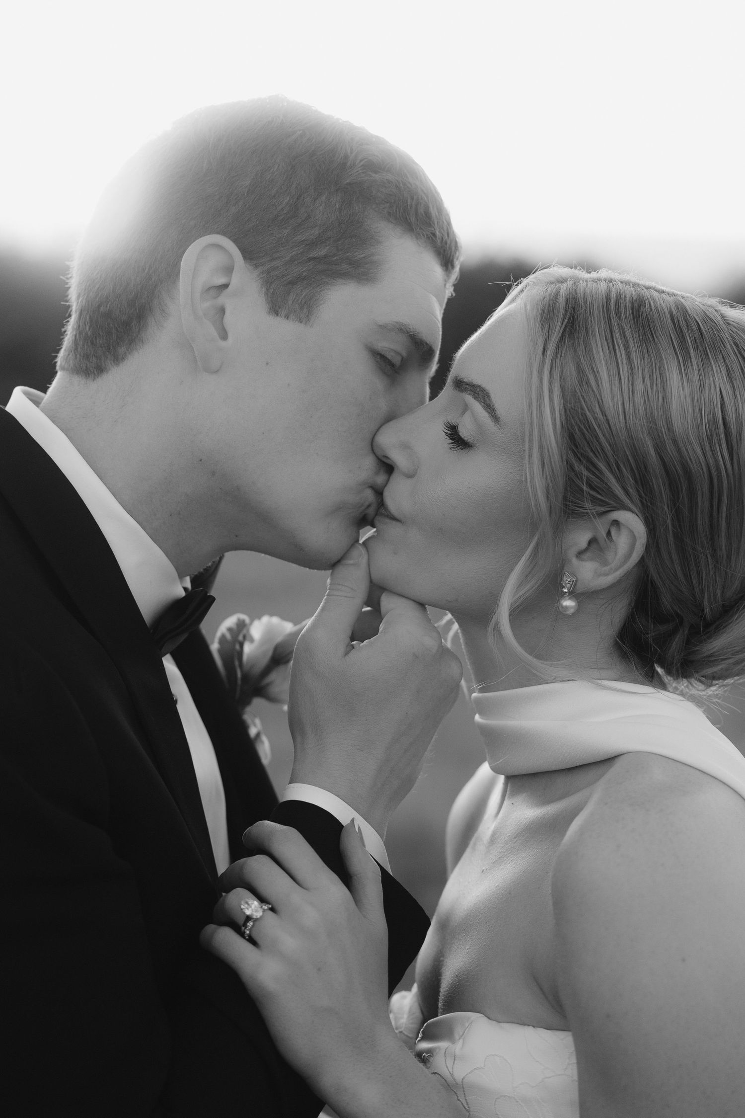 Bride and groom share a romantic kiss in black and white wedding portrait with soft backlight.