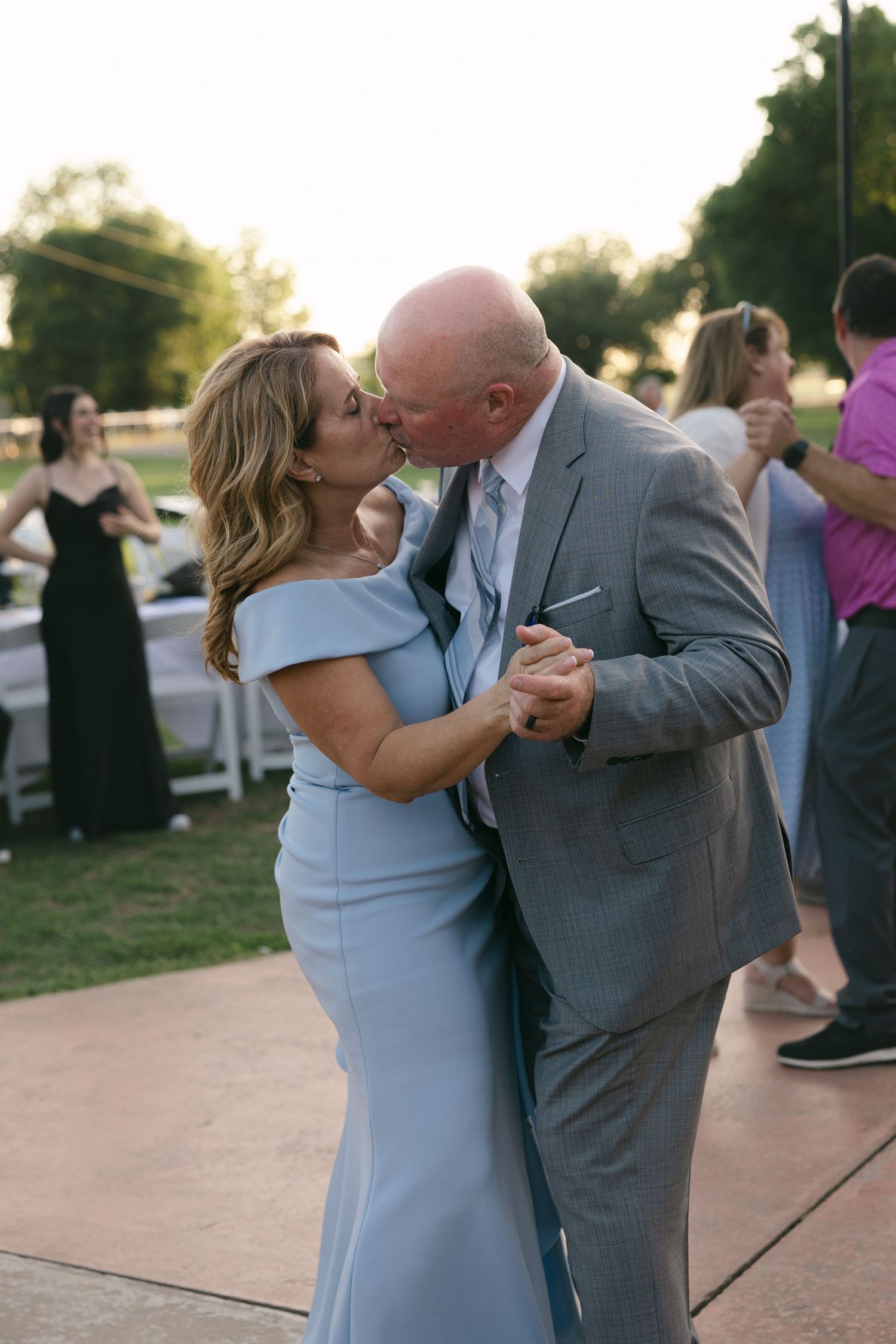 Couple sharing a kiss while dancing outdoors at a wedding reception, wearing light blue dress and gray suit.