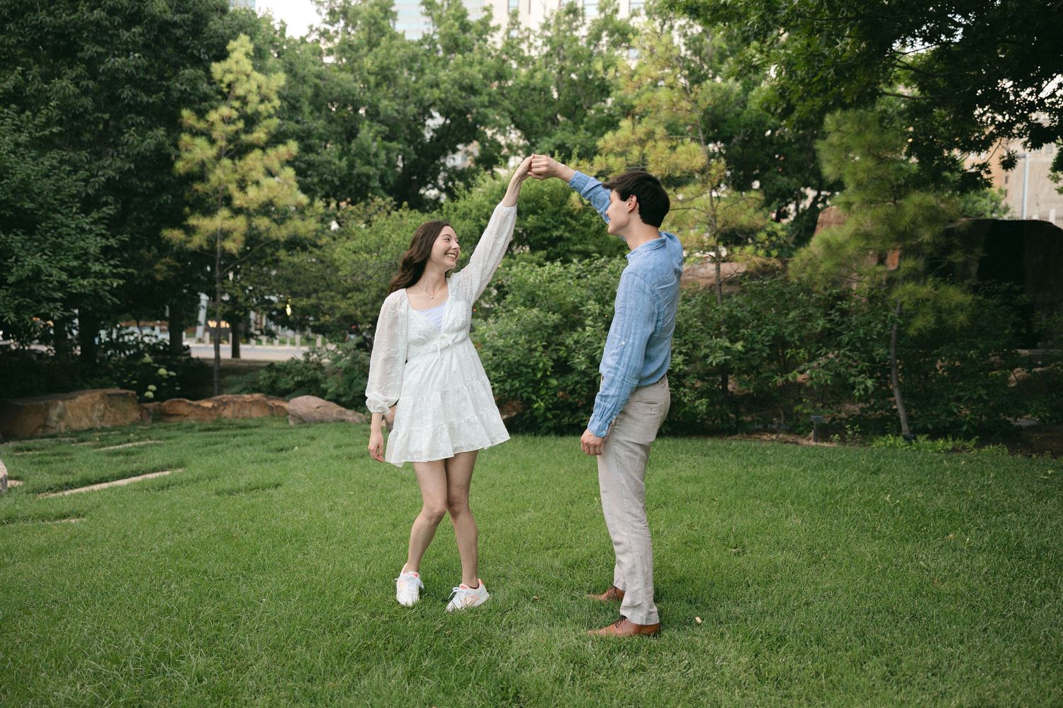 A couple dances joyfully in a lush green park, the woman in a white dress and man in a blue shirt holding hands.