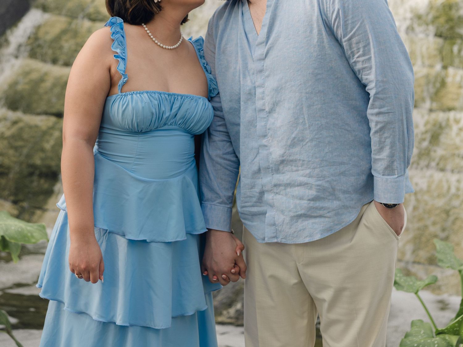 Couple in matching blue outfits holding hands outdoors, woman in tiered blue dress, man in light blue shirt and khaki pants.