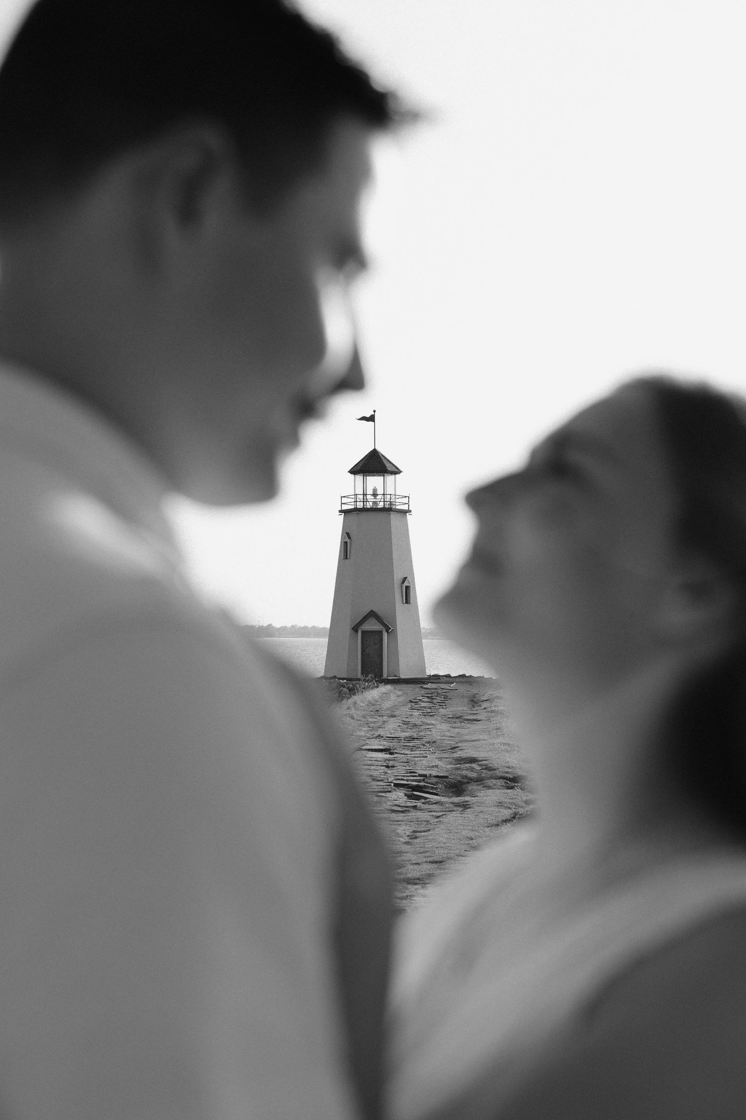 Black and white photo of a couple gazing at each other with a lighthouse reflected between them near the water.