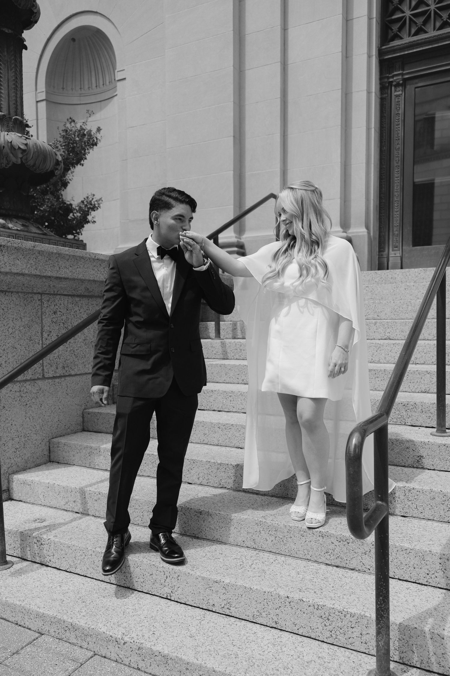 Black and white photo of a groom kissing his bride's hand on stone steps outside a grand building.