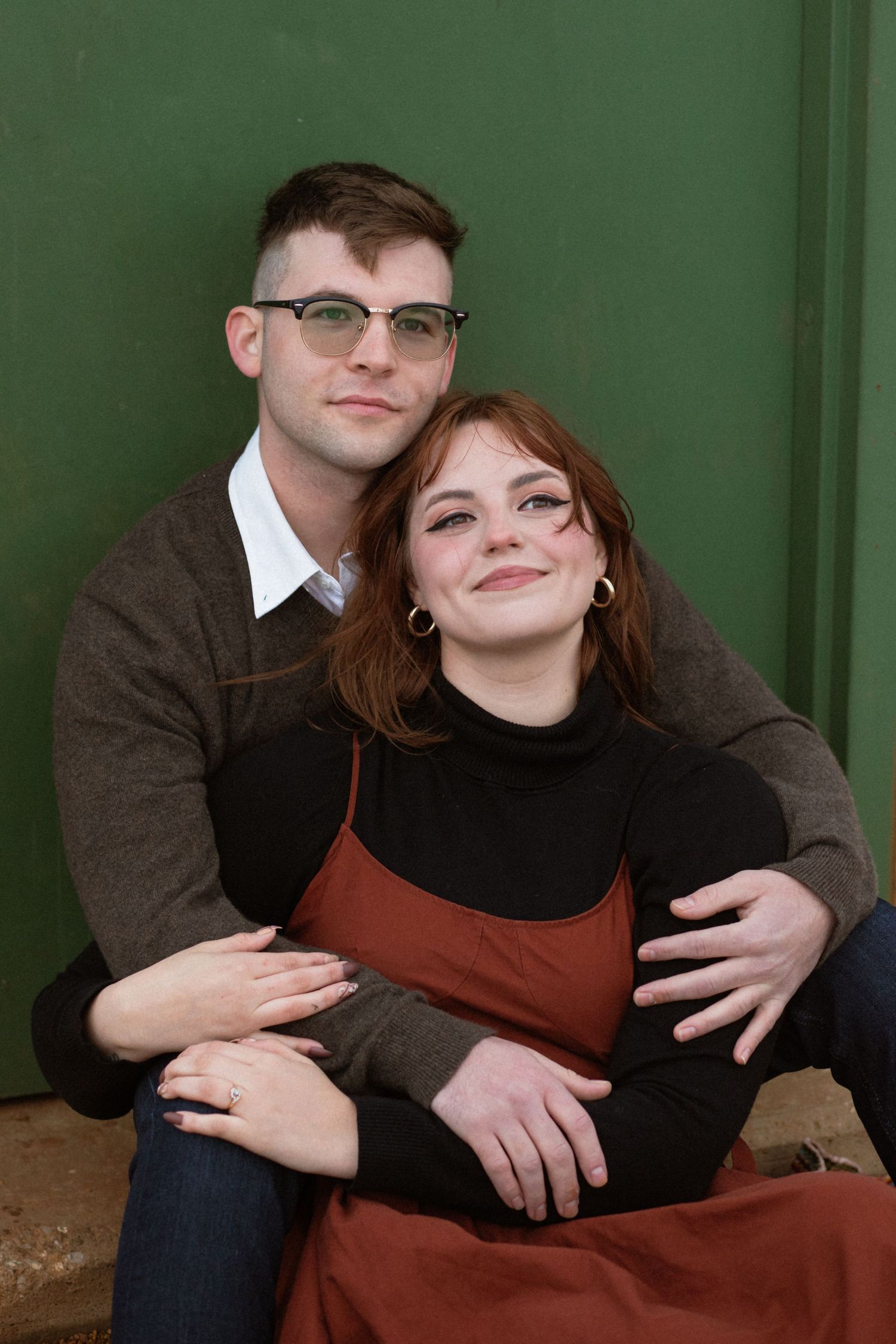 A couple sits close together against a green wall, wearing glasses and casual attire in earth tones.
