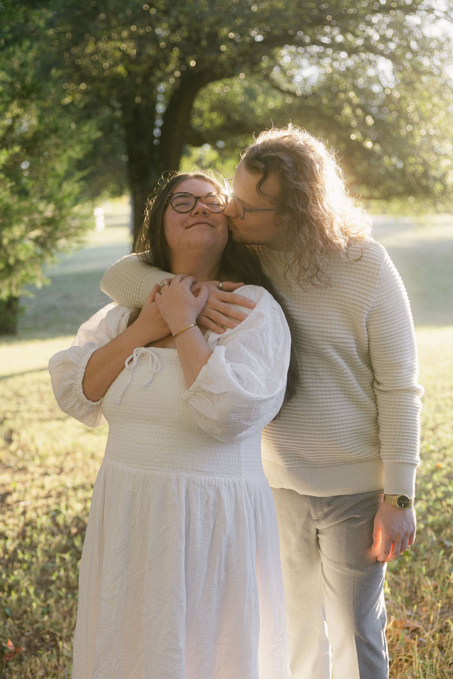 Two people share tender moments in a sunlit field wearing white and gray outfits.