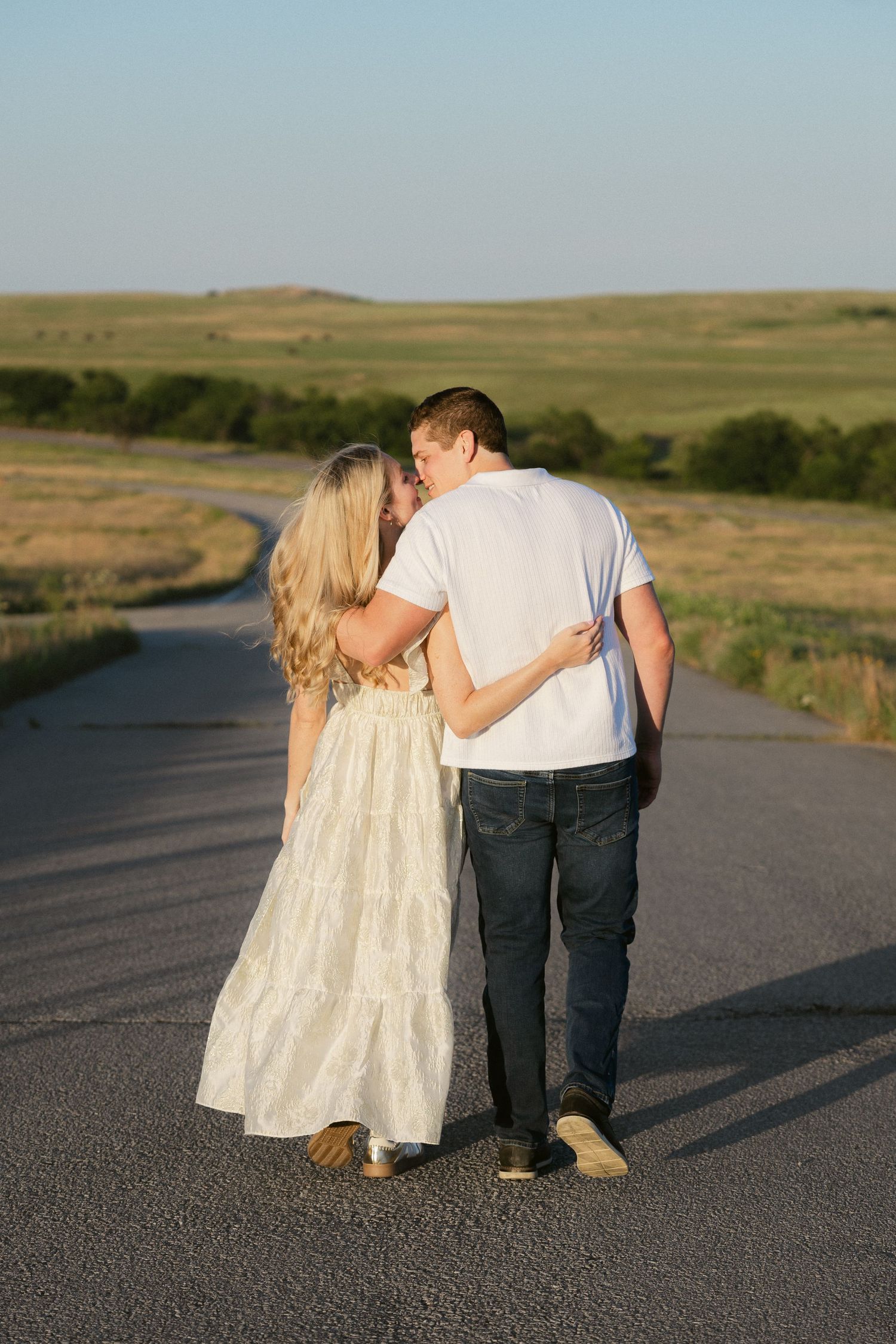 Two young children in light and dark outfits stand close together on a rural road with rolling hills in the background.