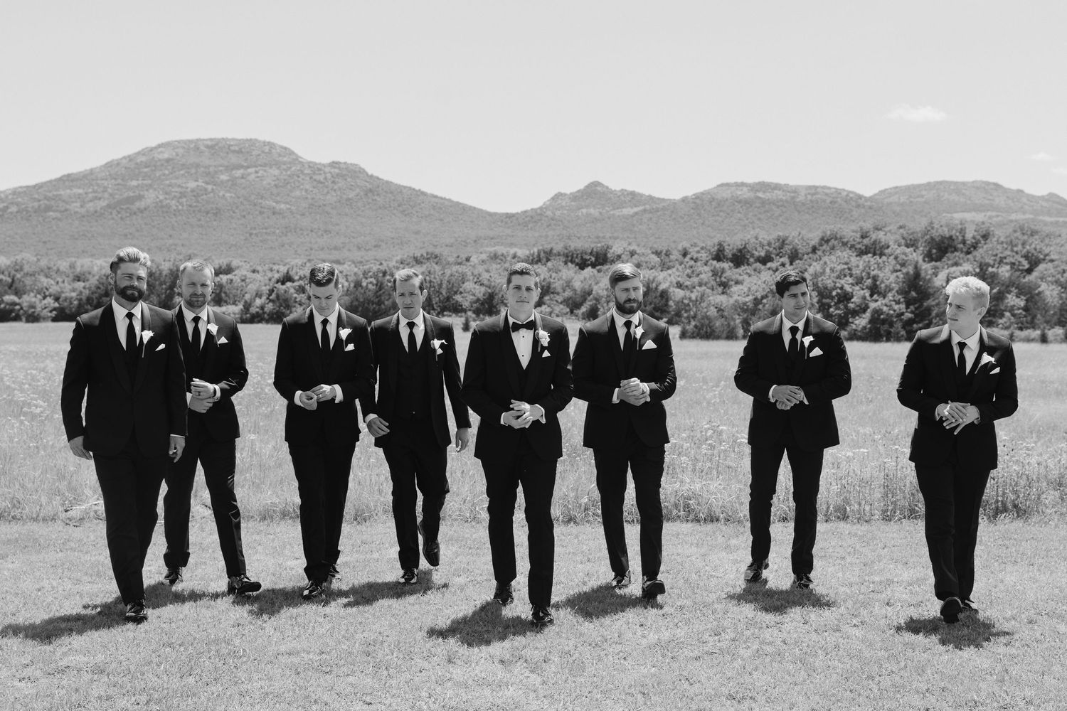 Seven groomsmen in black tuxedos walk together in a field with mountain backdrop in black and white wedding photo.
