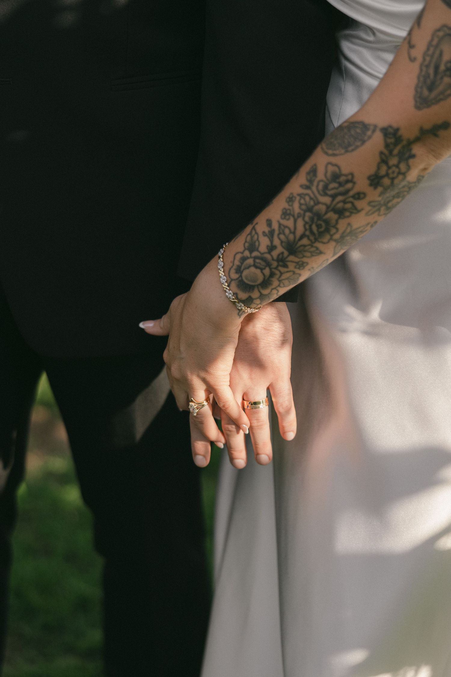 Close-up of newlyweds holding hands, showcasing wedding rings and bride's floral arm tattoo.