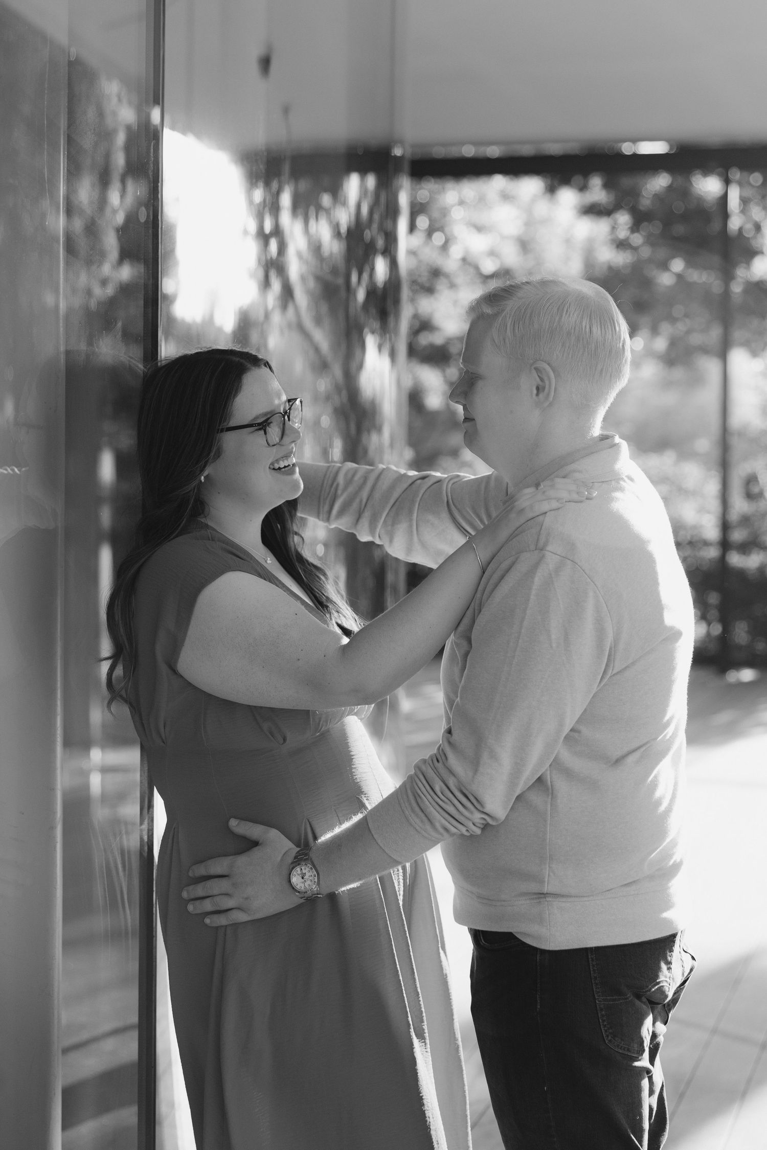 Black and white photo of a smiling pregnant couple embracing outdoors, woman in dress with glasses, man in casual shirt.