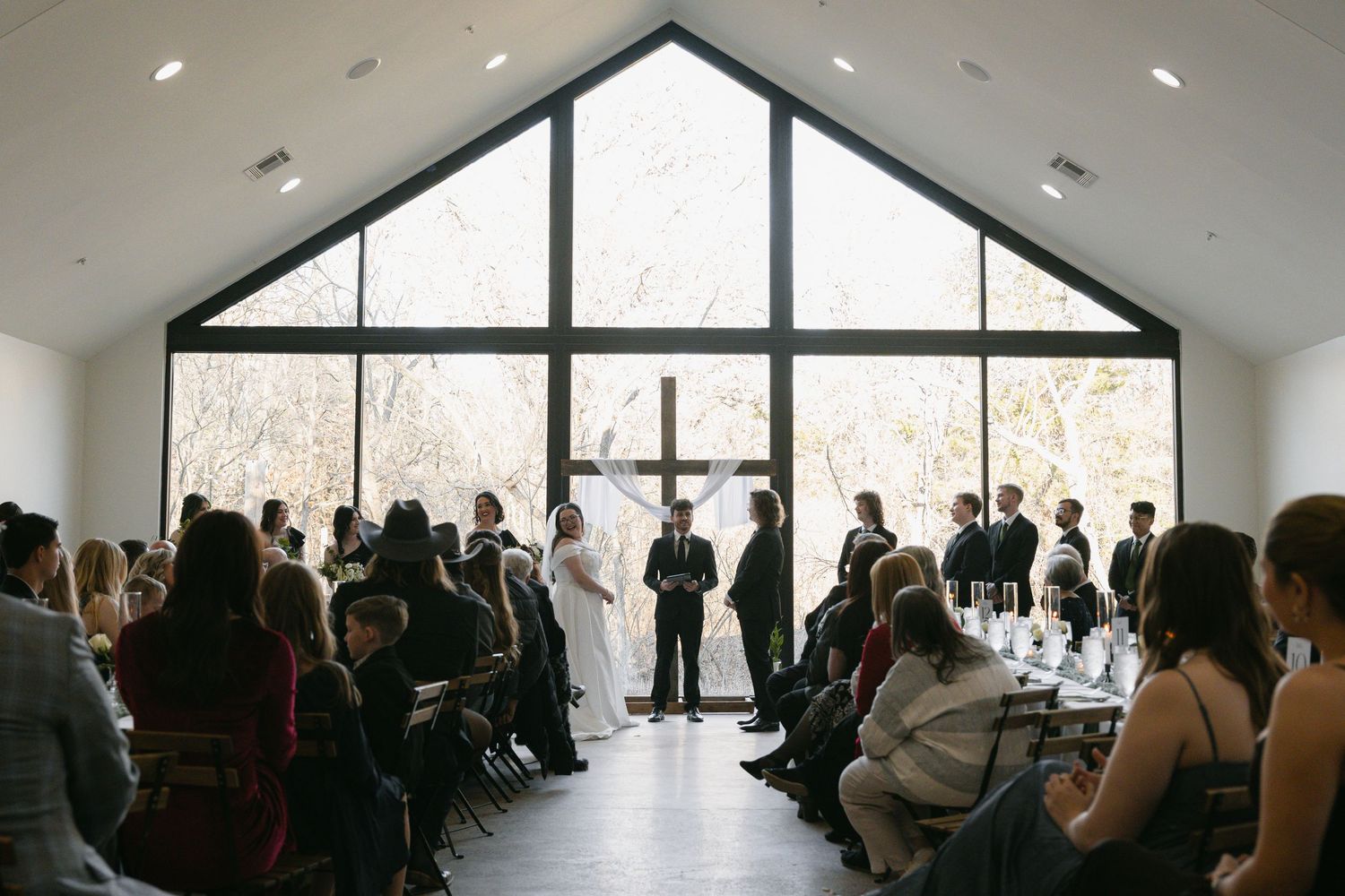 Wedding ceremony in a modern chapel with large windows, cross, and guests seated in rows.
