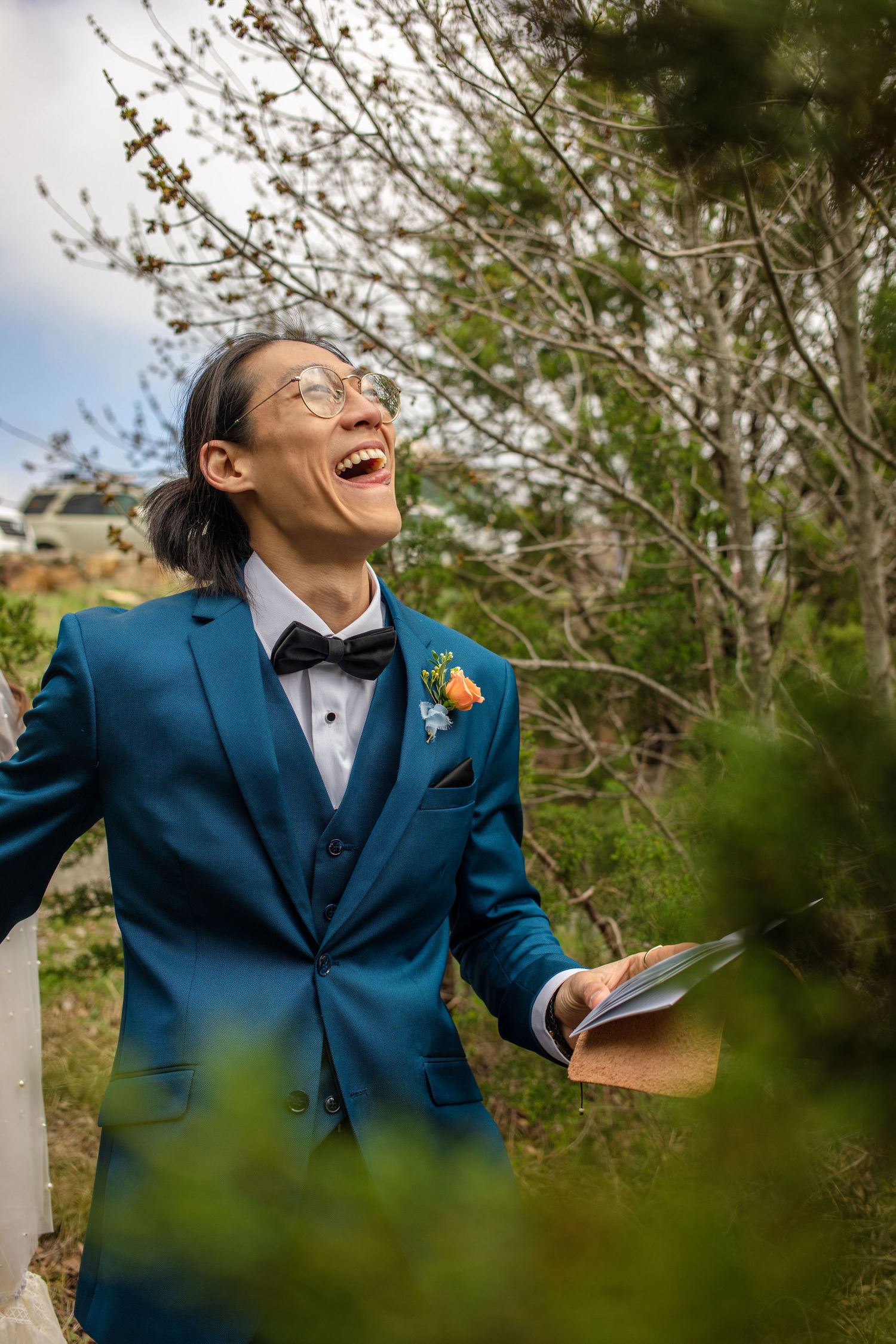 Wedding officiant in bright blue suit laughing joyfully while holding papers outdoors.