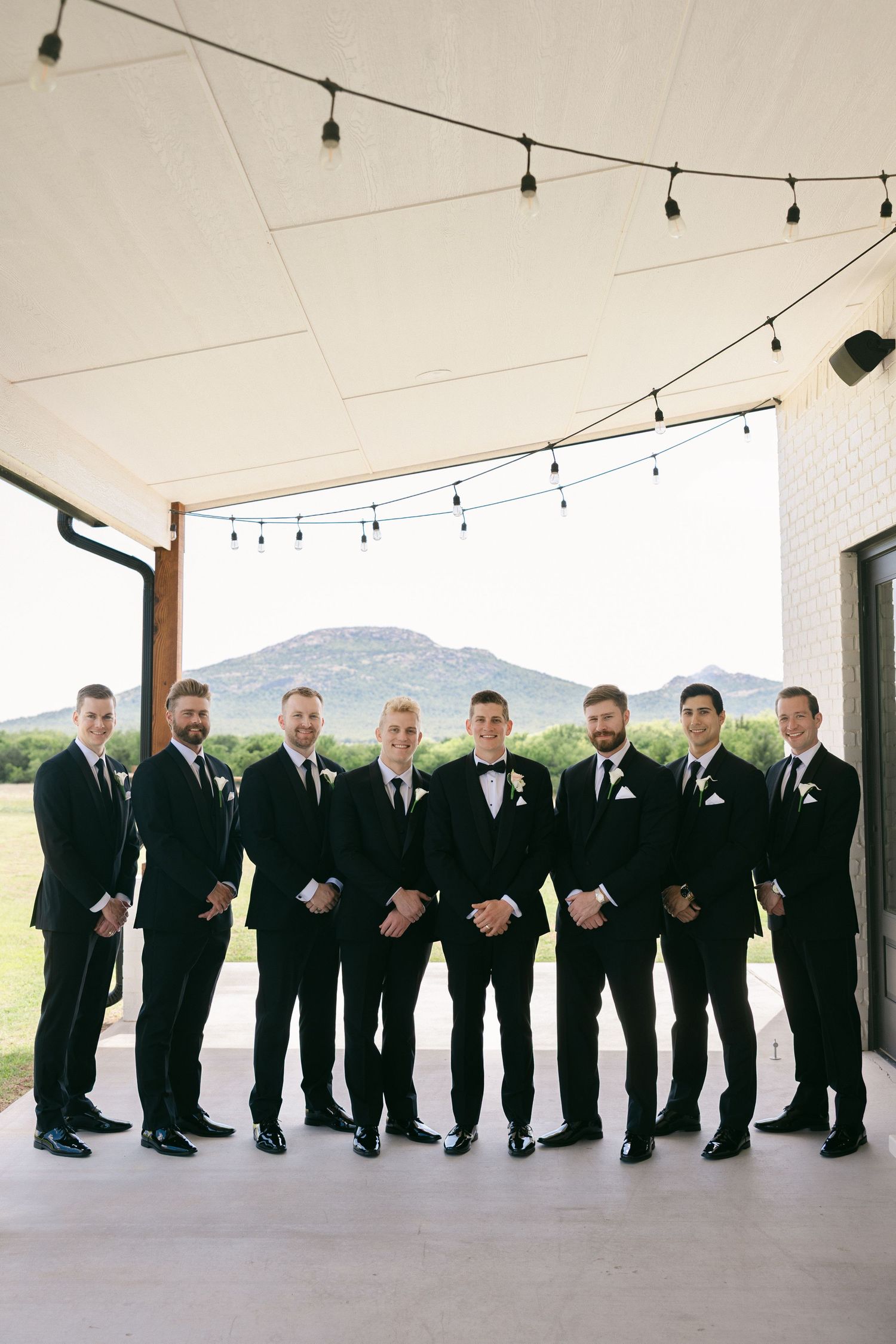 Eight groomsmen in black tuxedos pose together on a patio with string lights and mountain views.