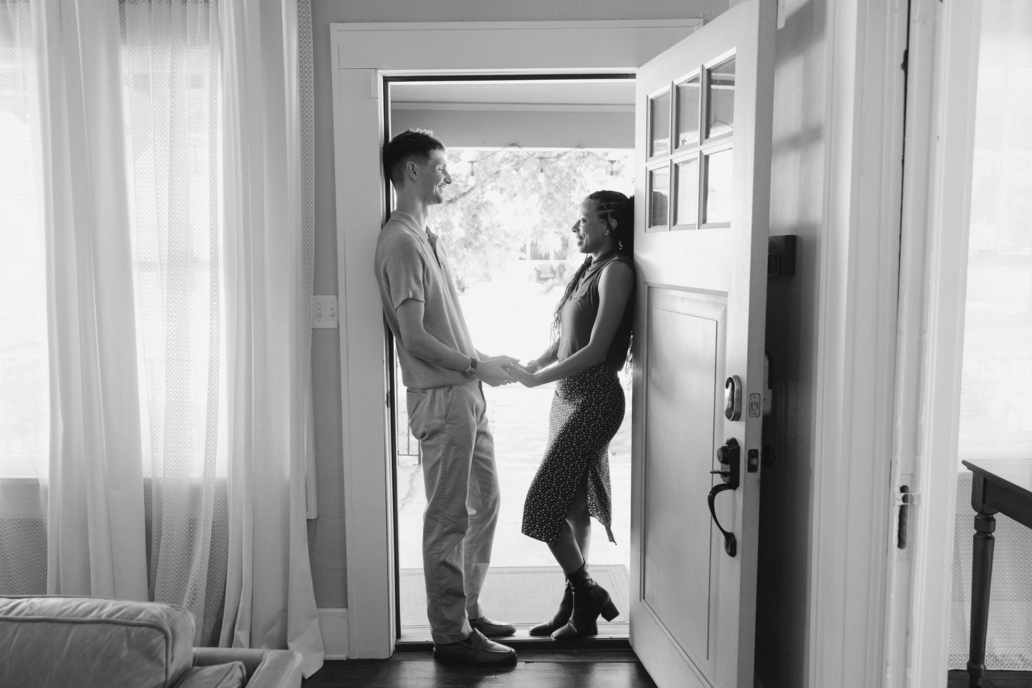 A couple holds hands while standing in an open doorway in a black and white romantic indoor photo.