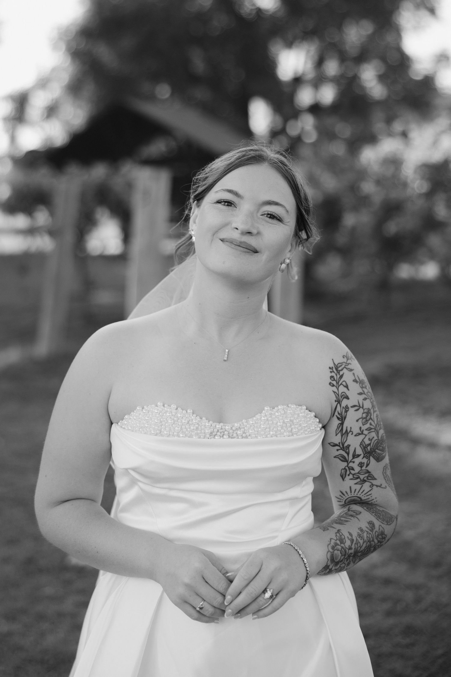 Smiling bride in strapless pearl-detailed gown with floral sleeve tattoo poses outdoors.