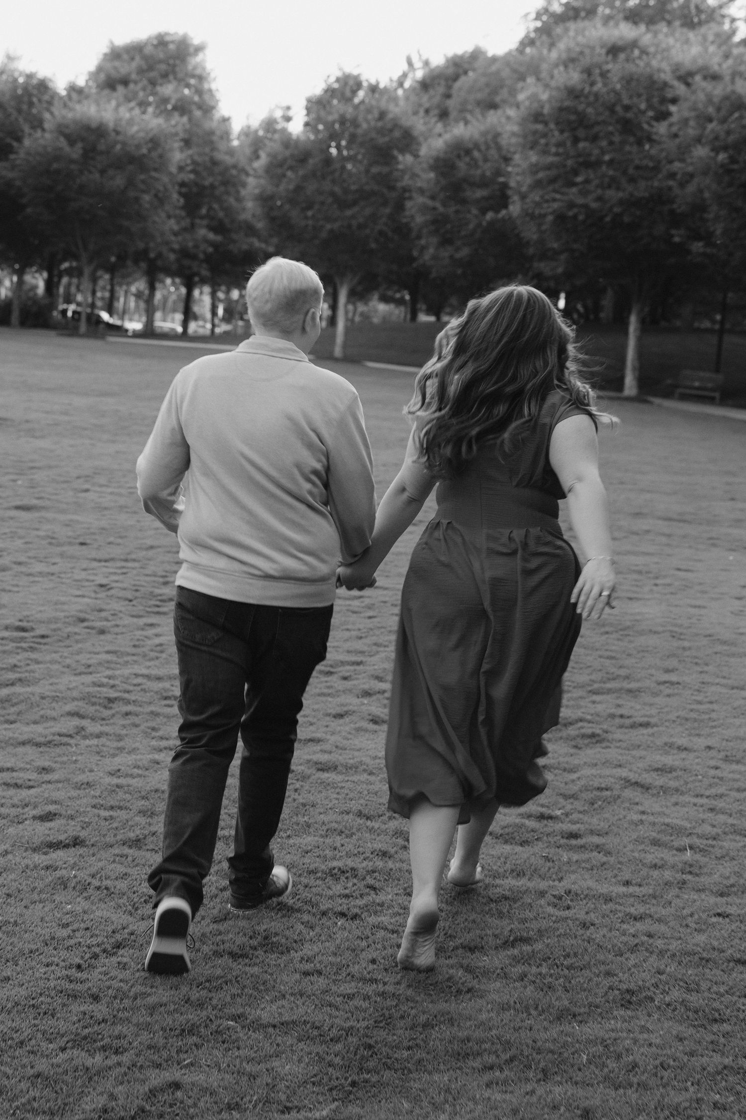 Black and white photo of a couple holding hands while walking across a grassy park lawn.