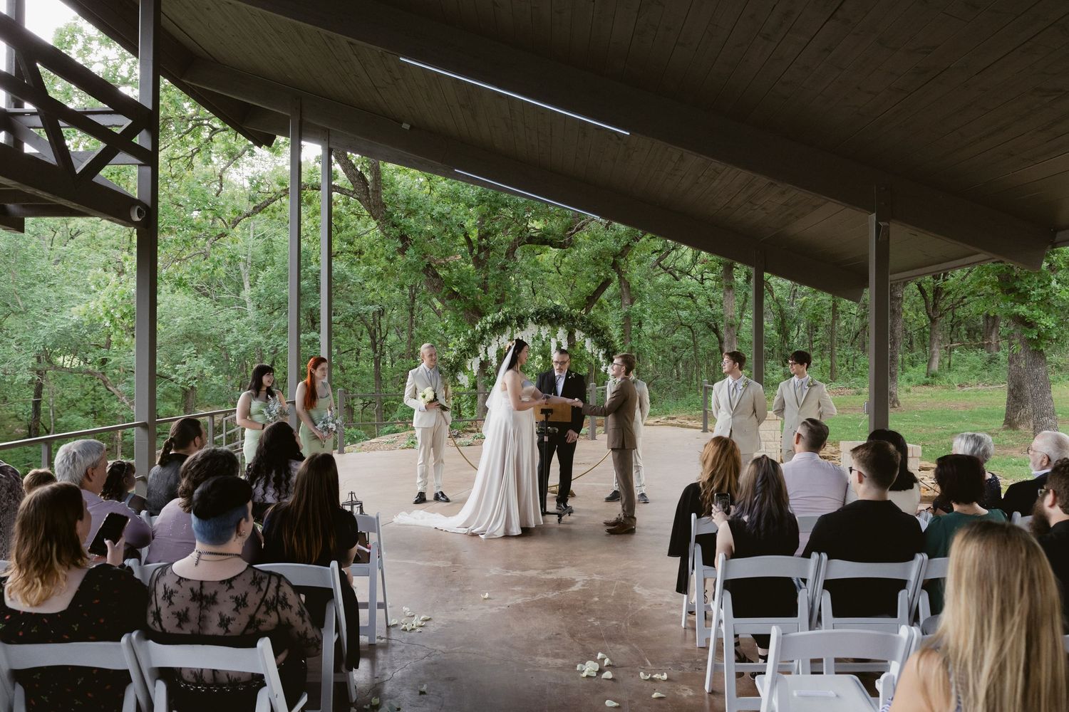 An outdoor wedding ceremony under a covered pavilion with white chairs and greenery in the background.
