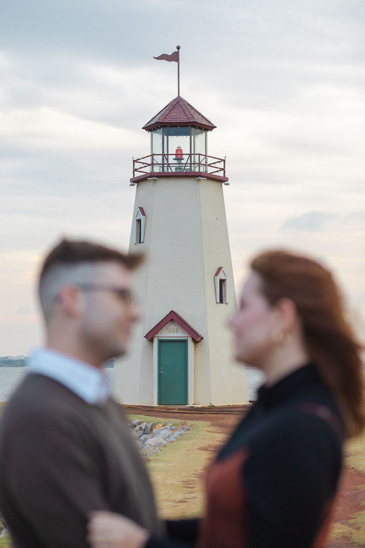 A lighthouse with a red roof and green door stands tall against a cloudy sky, with two people in soft focus in the foreground.