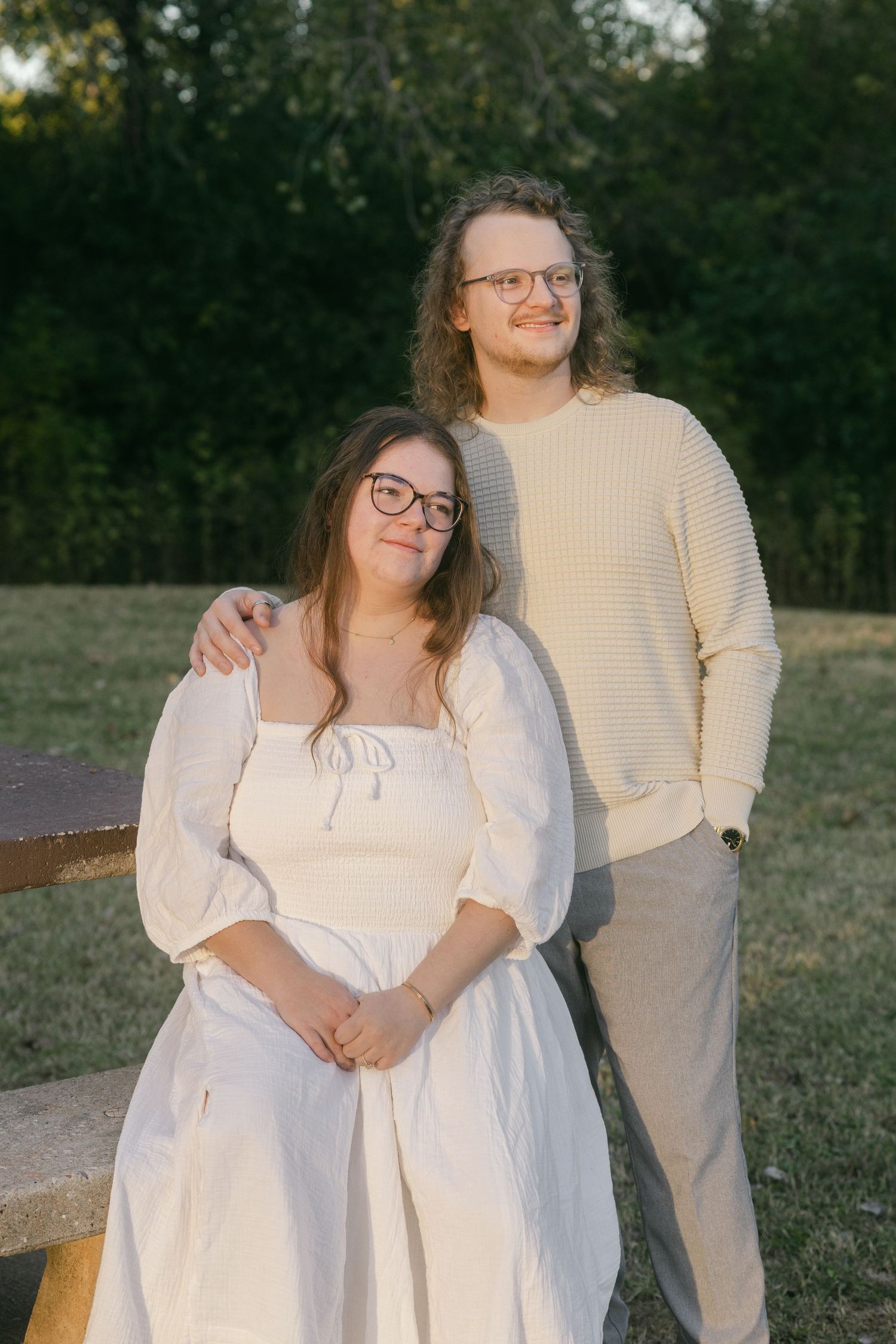 A couple in coordinating cream-colored outfits pose together outdoors during golden hour at sunset.