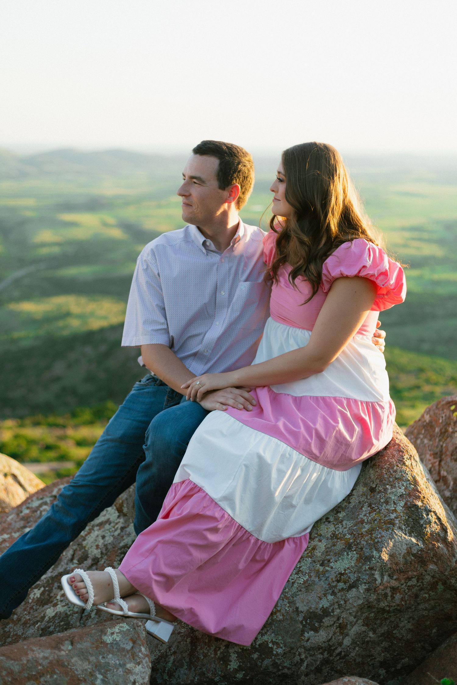 A couple sits together on a rocky overlook with scenic rolling hills stretching into the distance at sunset.