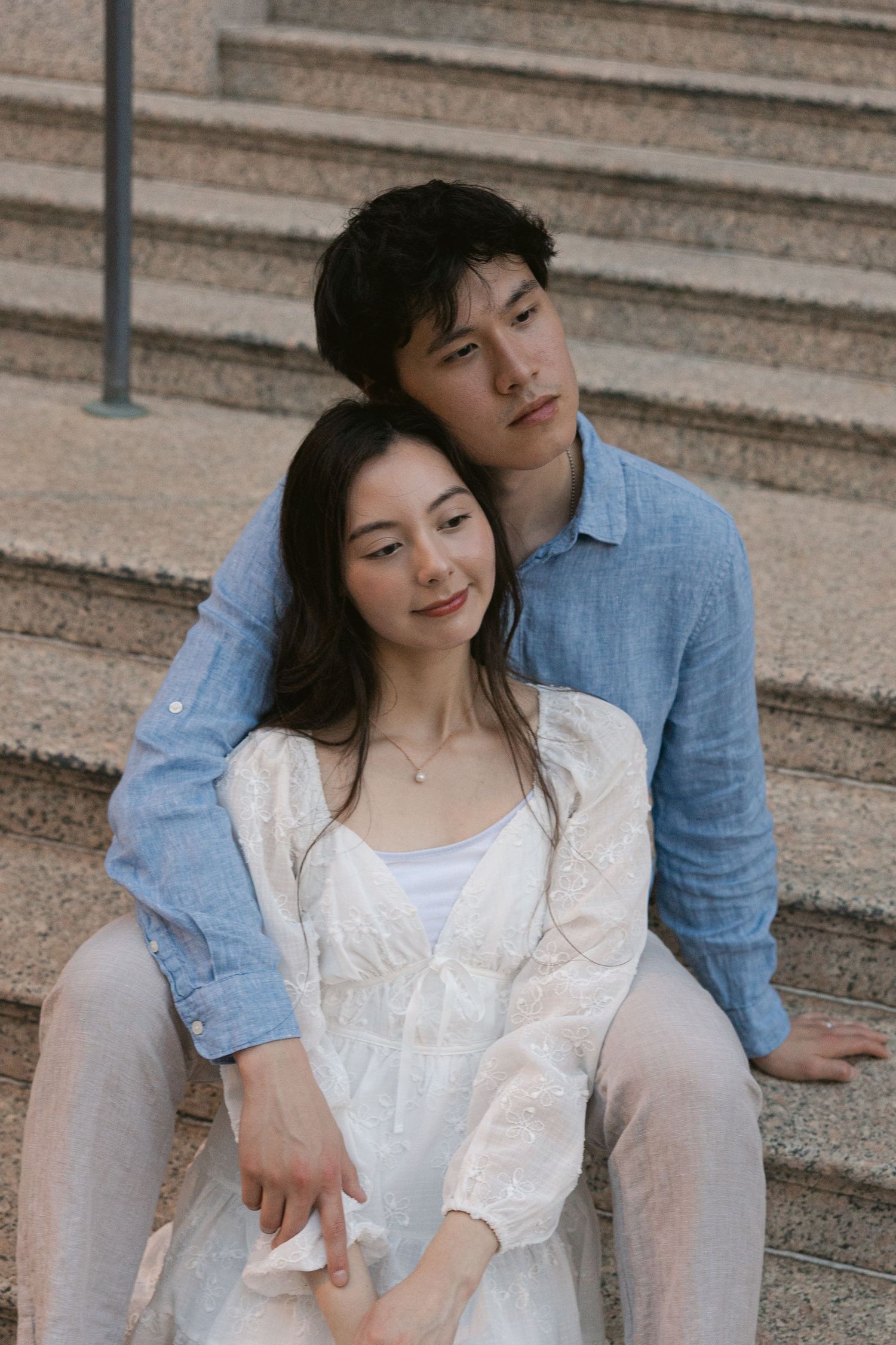 A couple sits on stone steps, woman in white dress leaning on man in blue shirt, in a tender romantic pose.