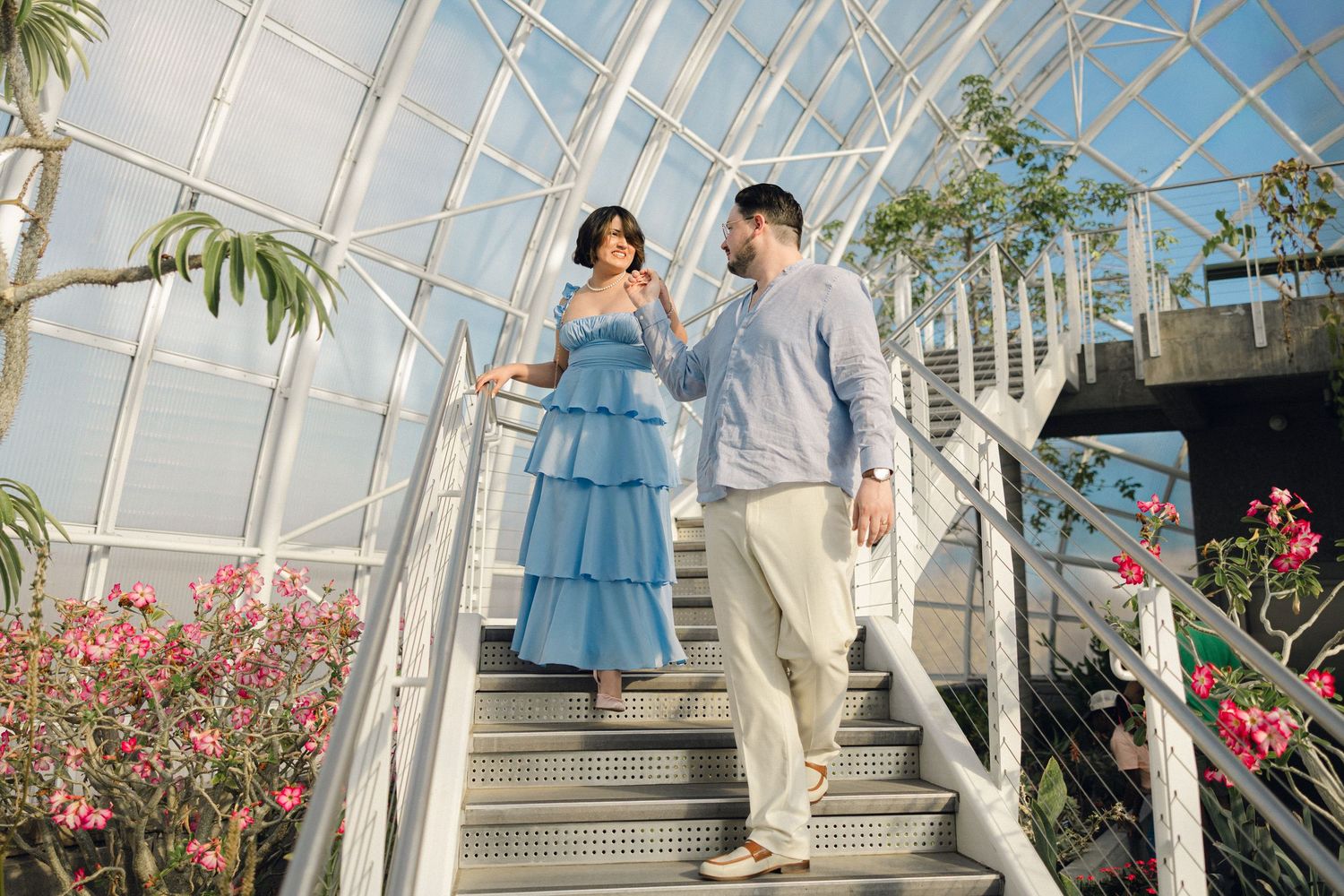 Couple in elegant attire walking down stairs in a greenhouse with white railings and tropical flowers.