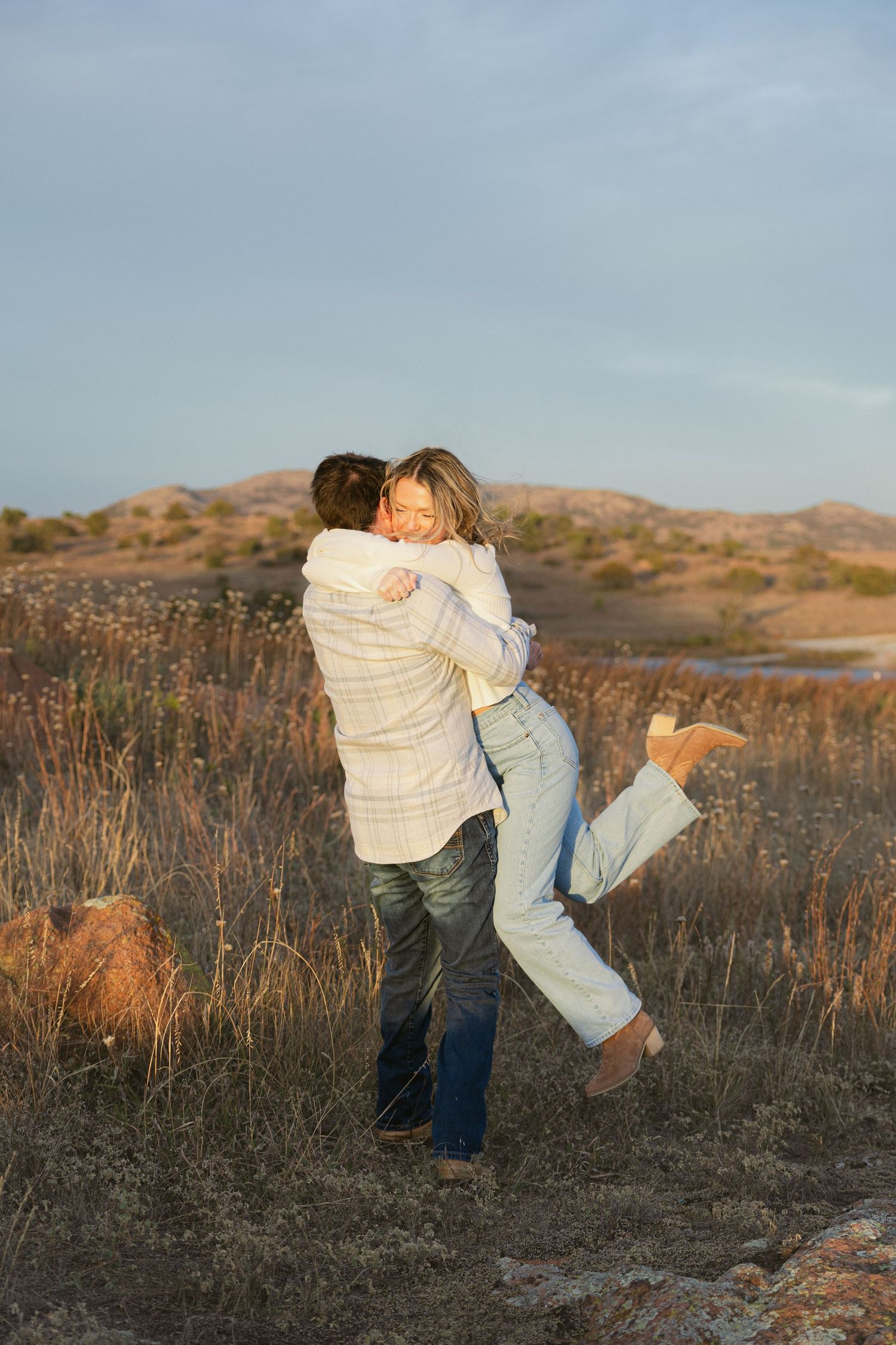 A couple embraces in a romantic moment at sunset against a backdrop of golden hills and dry brush.
