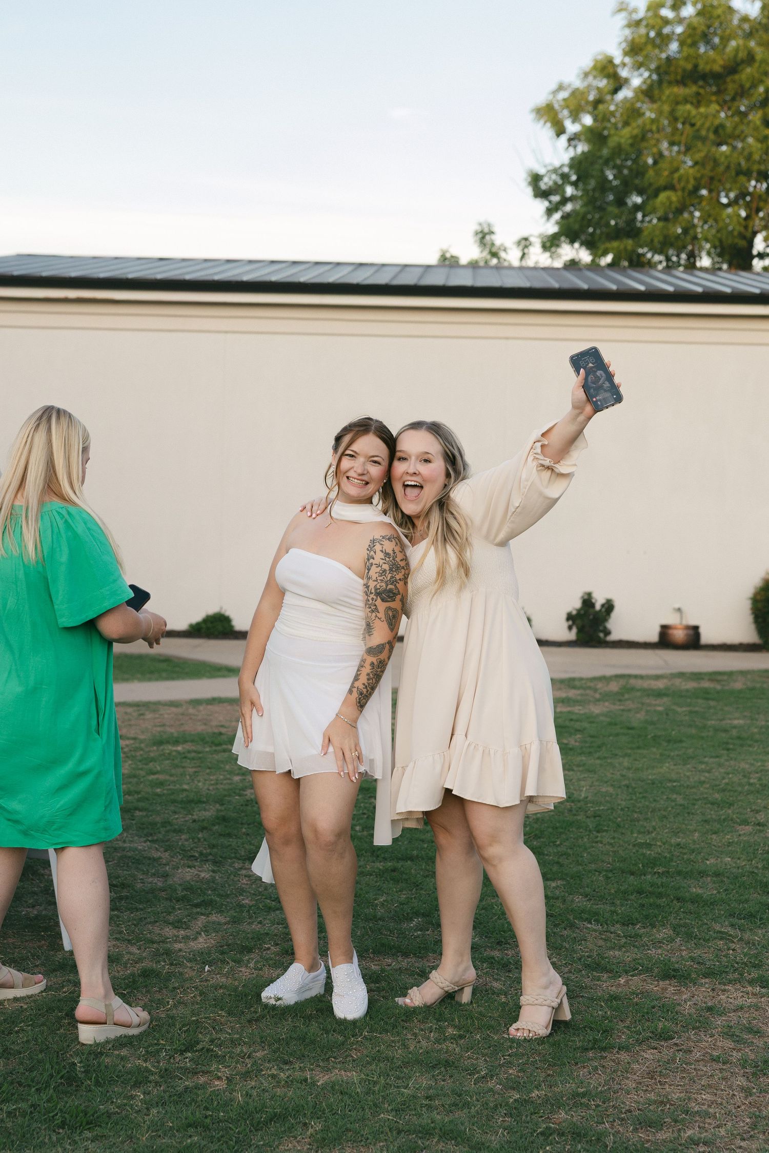 Two women in white dresses take a selfie outdoors on green grass, smiling, while another woman in green stands nearby.