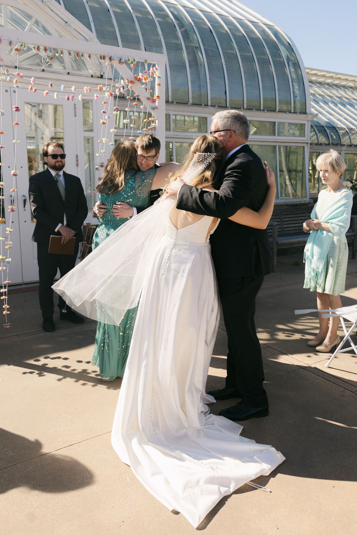 Bride hugging family members outside a glass greenhouse venue during a wedding ceremony on a sunny day.